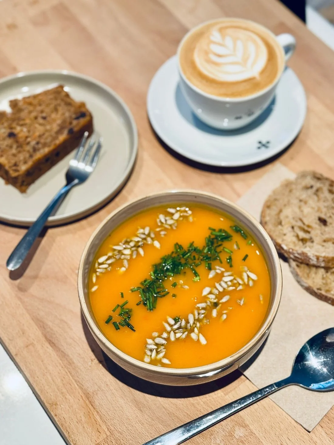 Soupe de citrouille garnie de graines et d'herbes, accompagnée de pain, avec un cappuccino avec un latte art et un cake à la carotte sur une table en bois.