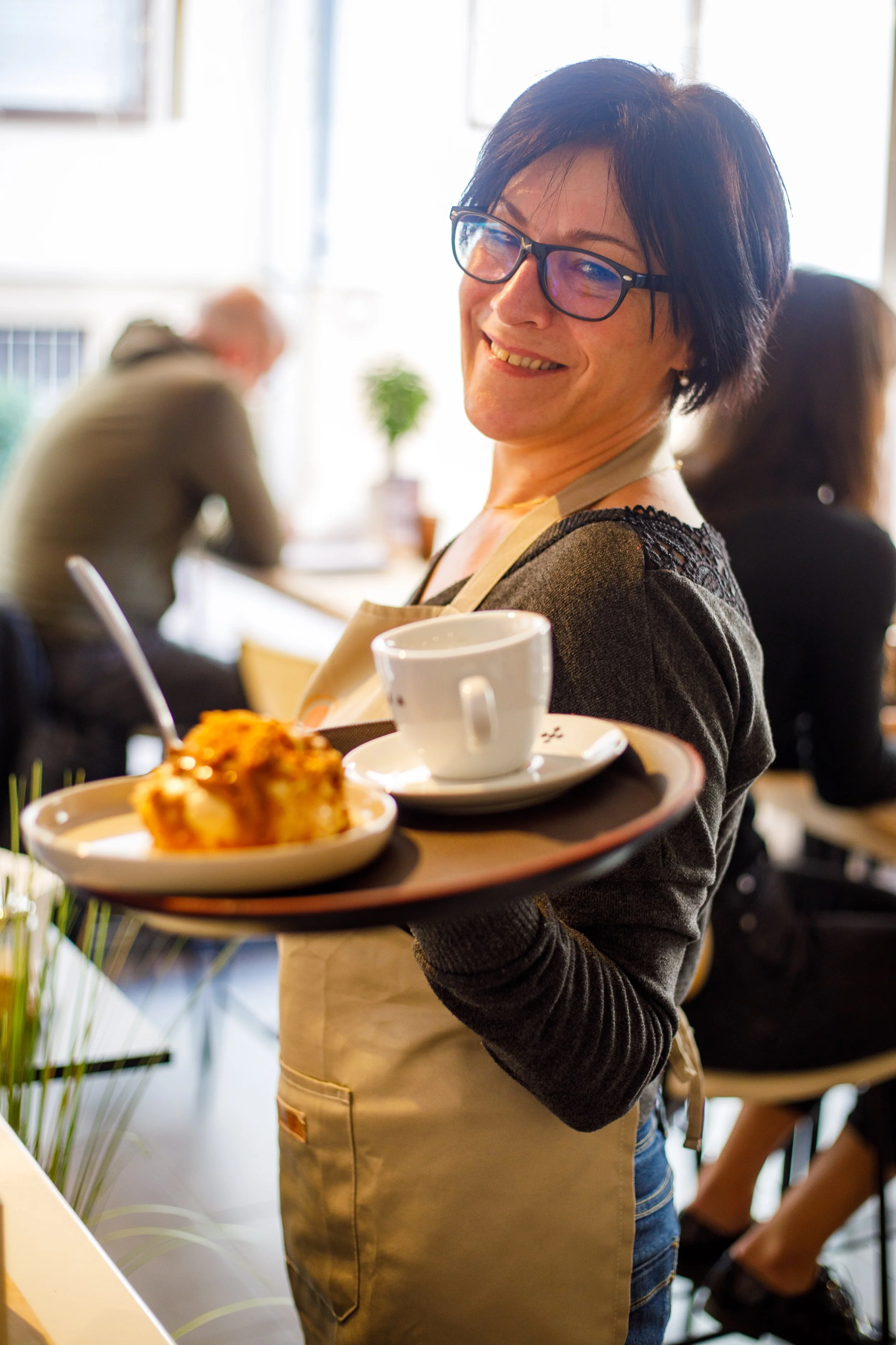 Une serveuse souriante portant un plateau avec une tasse à café et un dessert dans un café.
