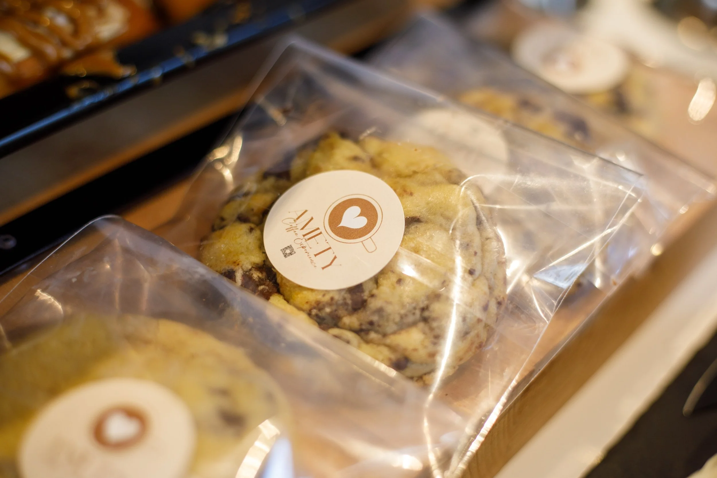Gâteau aux pépites de chocolat dans un emballage en plastique à rabat, avec un étiquette ronde indiquant 'AMITY' et un cœur dans une tasse, posé sur une surface en bois.
