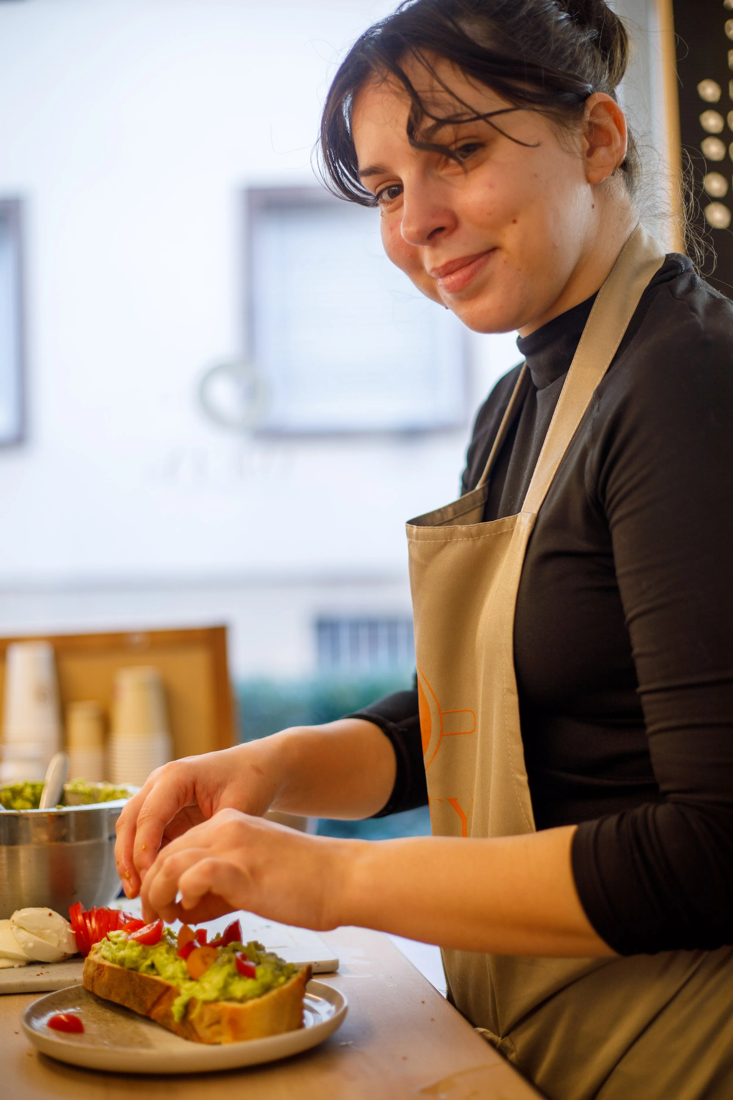 Une femme en tablier préparant un sandwich avec de la guacamole et des légumes dans une cuisine.