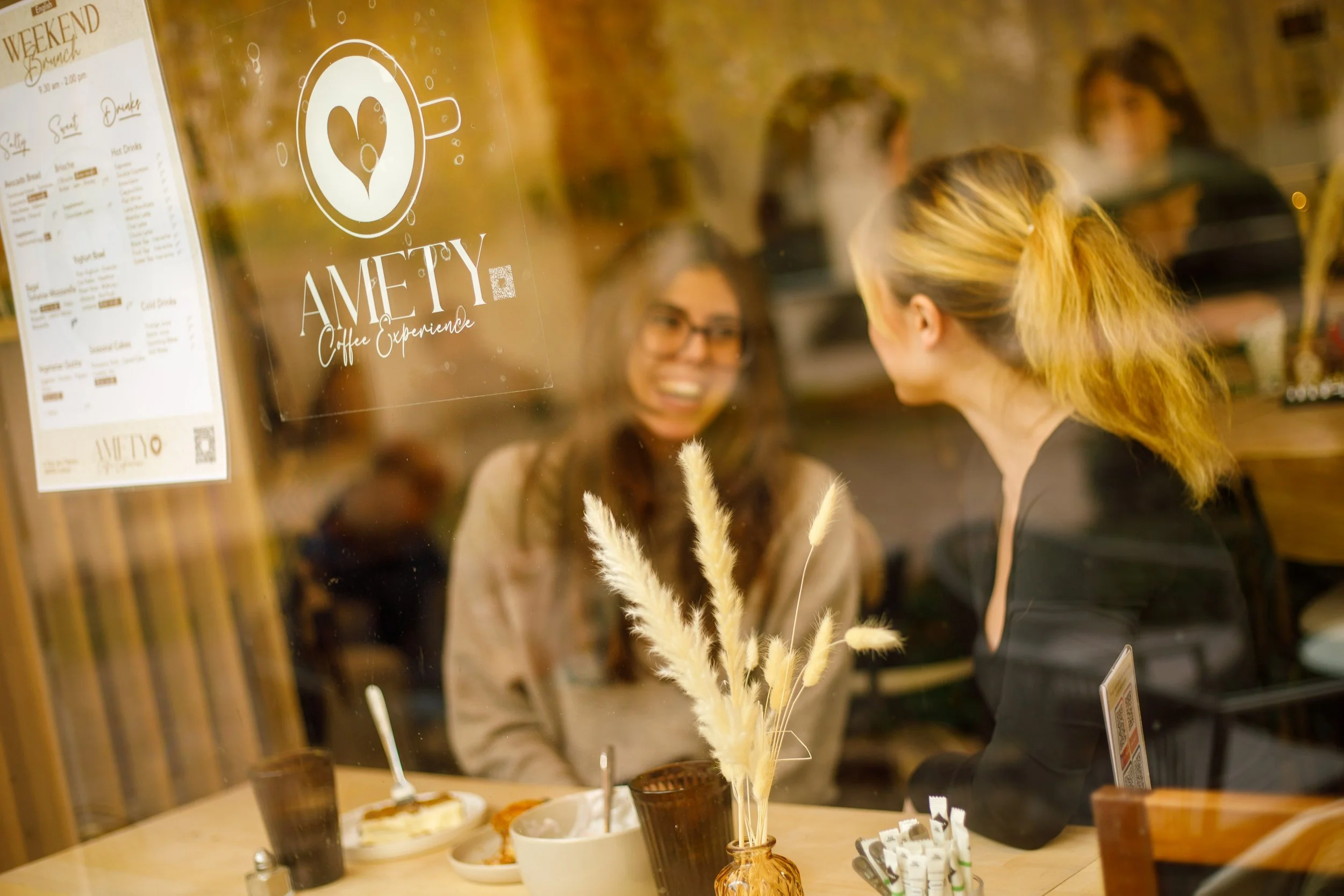 Deux femmes discutent dans un café, vue à travers une vitre avec le logo "AMETY Coffee Experience"