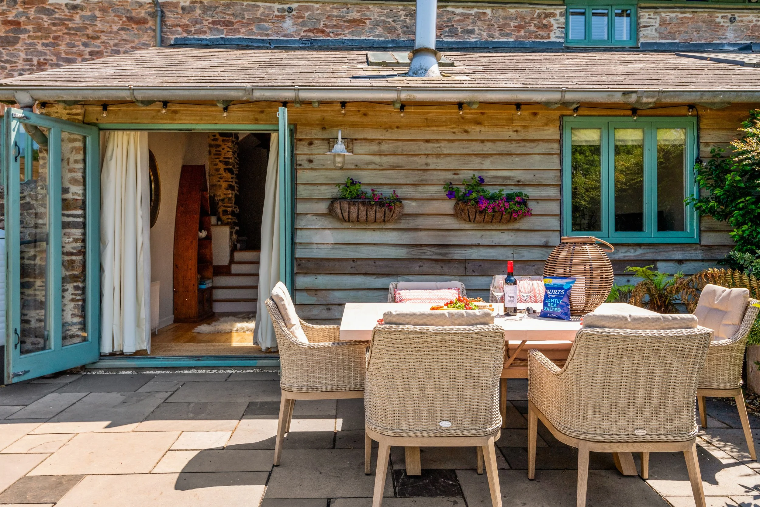 Outdoor patio with a rectangular table surrounded by six wicker chairs, set with a bottle of red wine, a wine glass, a package of salted crackers, and a woven decorative item, against a rustic wooden house with open blue doors, flower baskets, and a window.