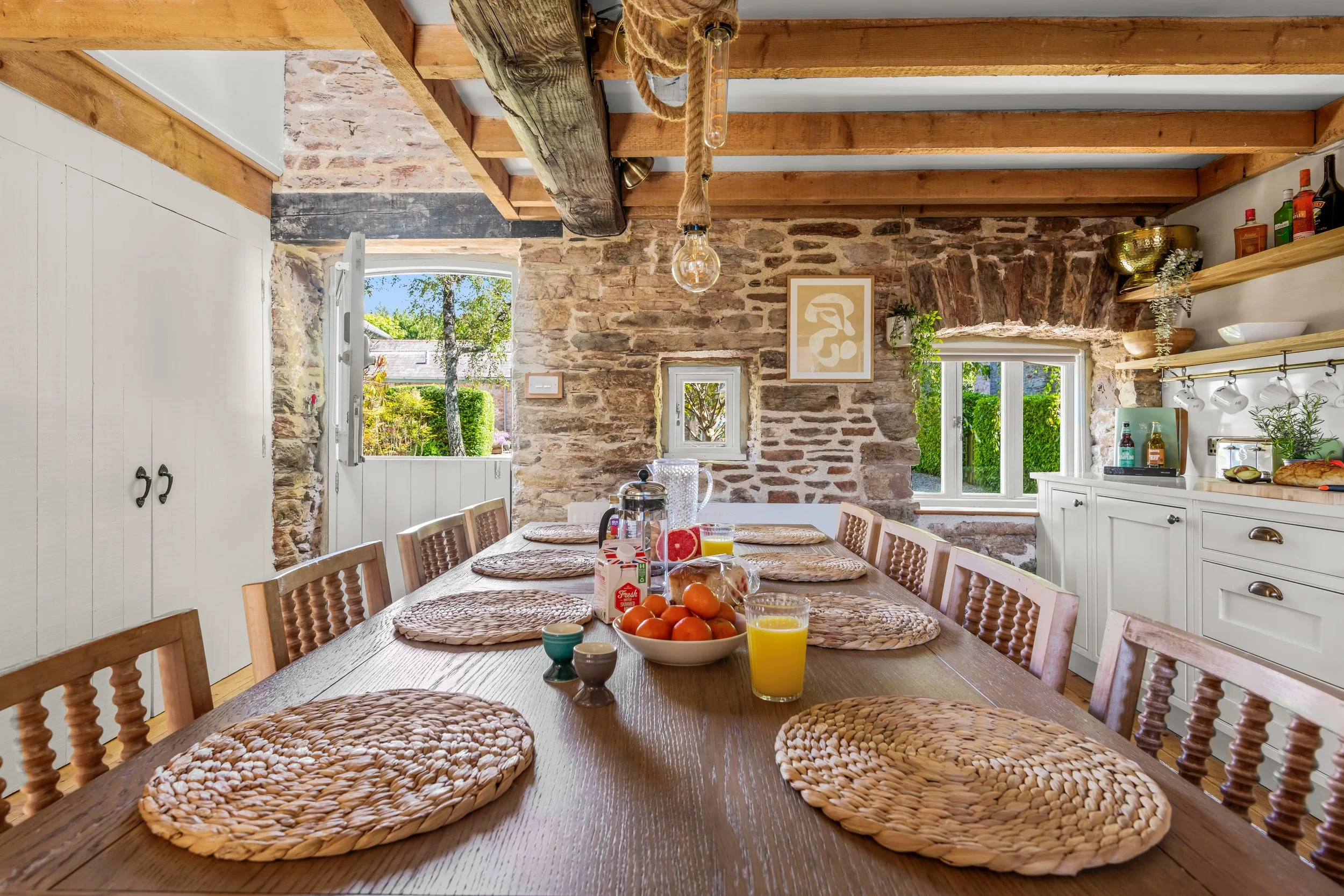 A rustic dining room with a long wooden table set with woven placemats, a bowl of oranges, glasses of orange juice, and breakfast items. The room has stone walls, exposed wooden beams, and a window showing a garden outside.