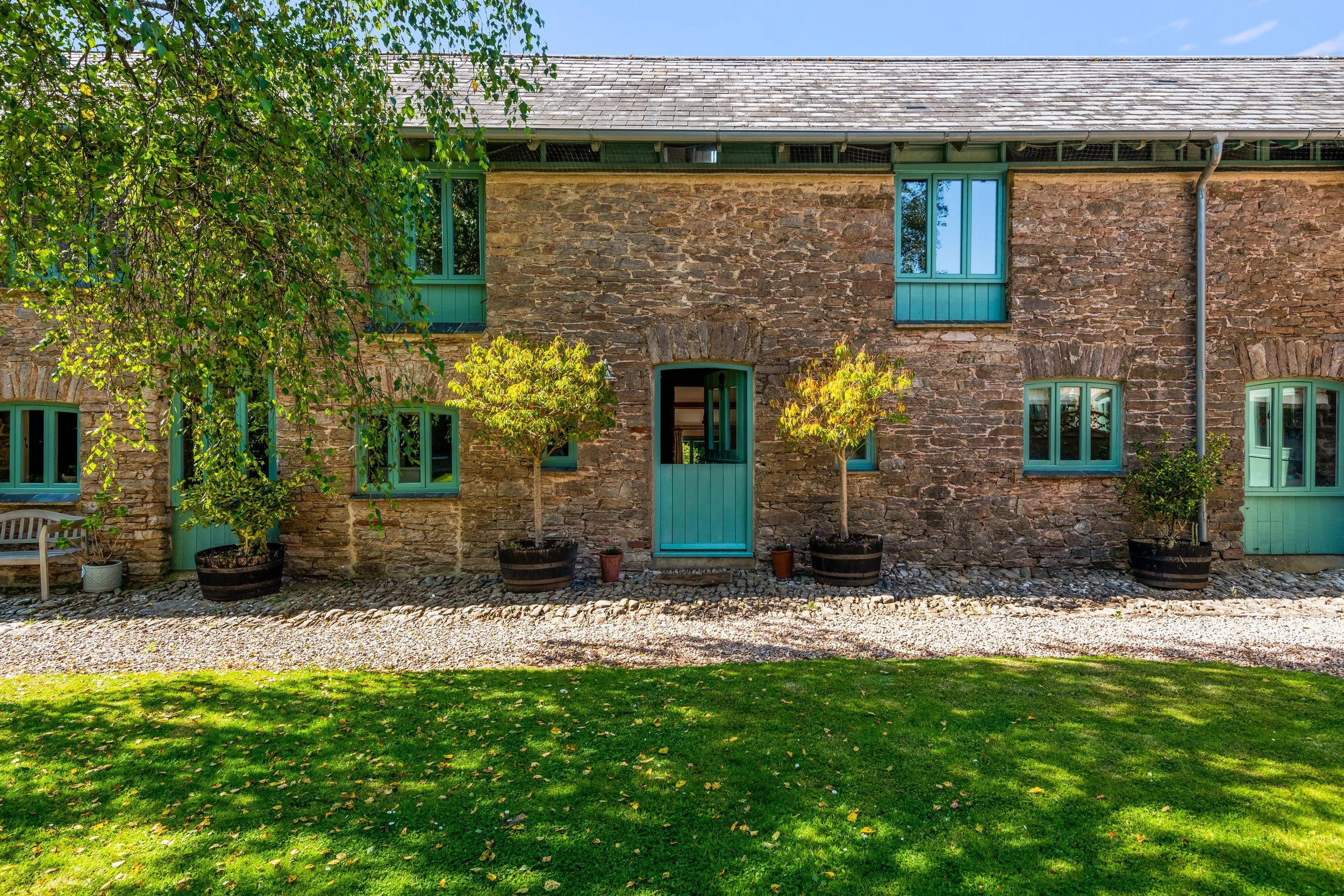 Stone house with green window frames and door, surrounded by potted trees and a grassy lawn.