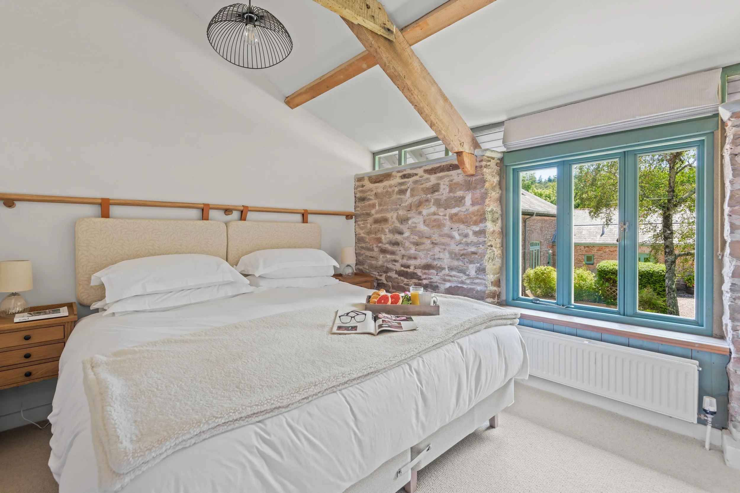 A cozy bedroom with a large bed, cream-colored bedding, and a beige headboard. There are wooden nightstands with lamps on each side. An open book, glasses, and a tray of breakfast items including grapefruit, orange juice, and a cup are on the bed. Th