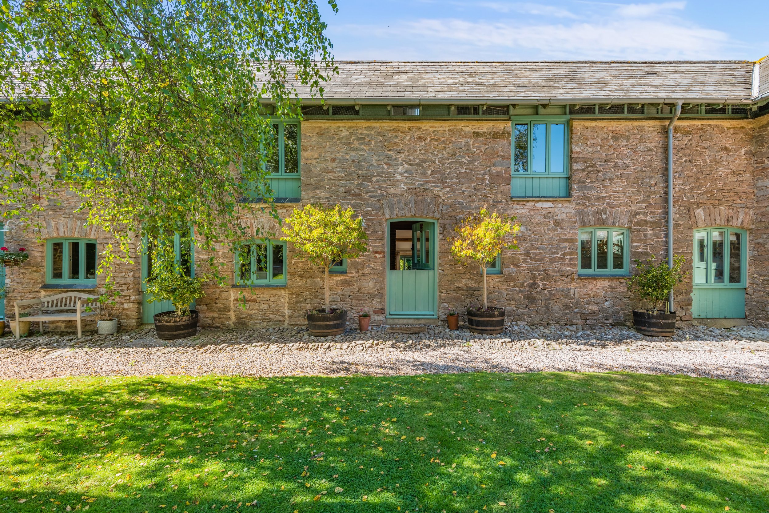 A rustic stone house with green window frames and a green door. There are small trees in pots along the front and a green lawn with some fallen leaves.