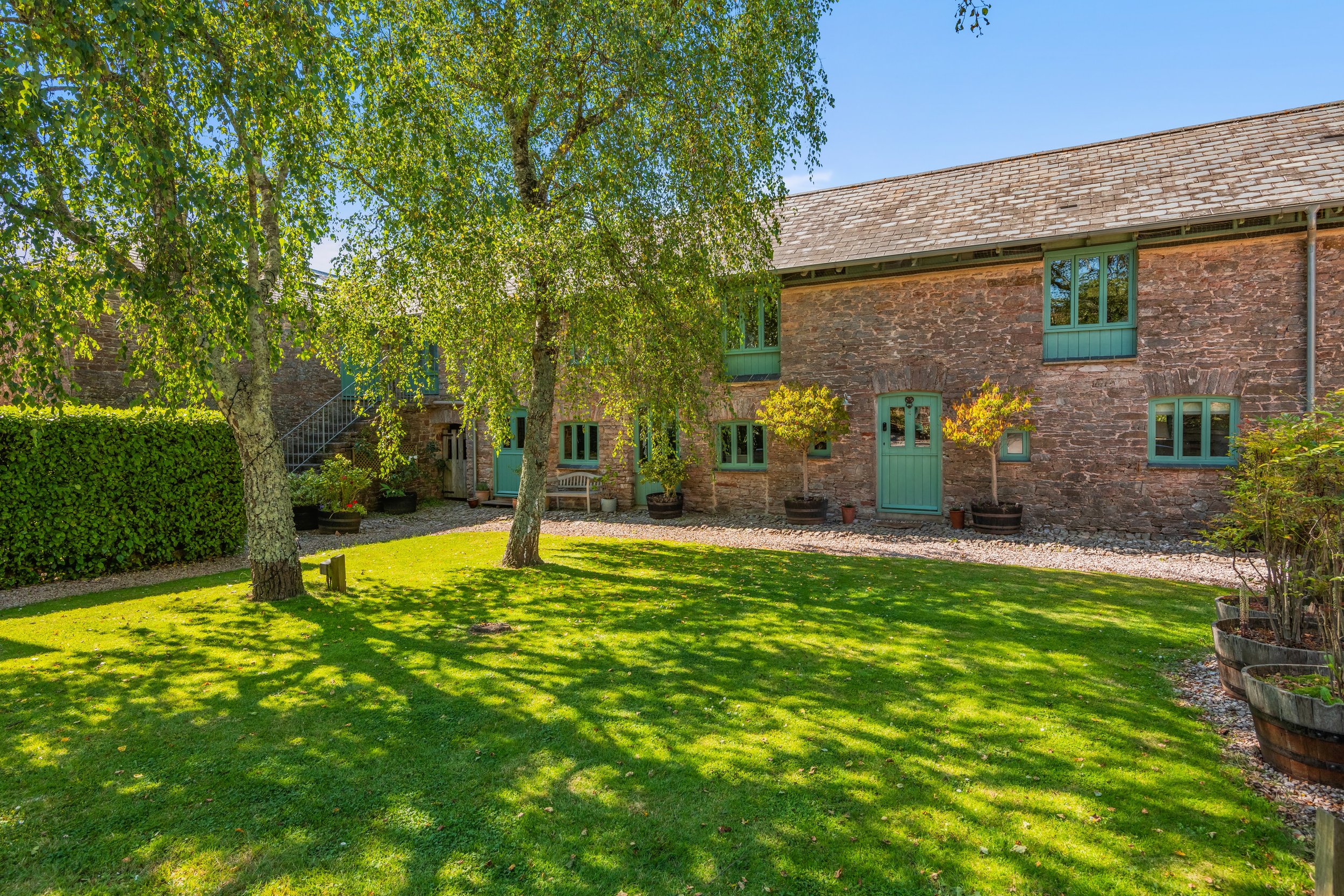 A peaceful yard with a lush green lawn, trees casting shadows, potted plants along the stone building wall, and a hedge on the left side. The building has a brick exterior with green trim around windows and door, and a sloped roof.
