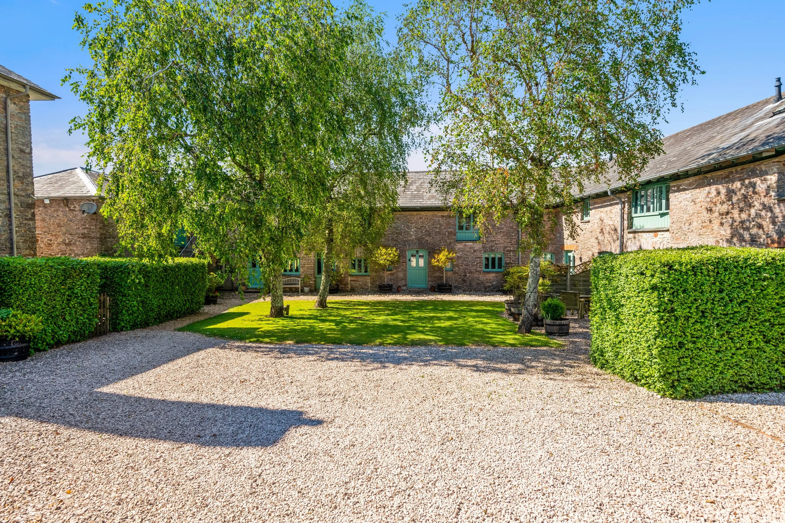 A courtyard with a gravel pathway, green grass, trees, and brick buildings with green windows and doors.