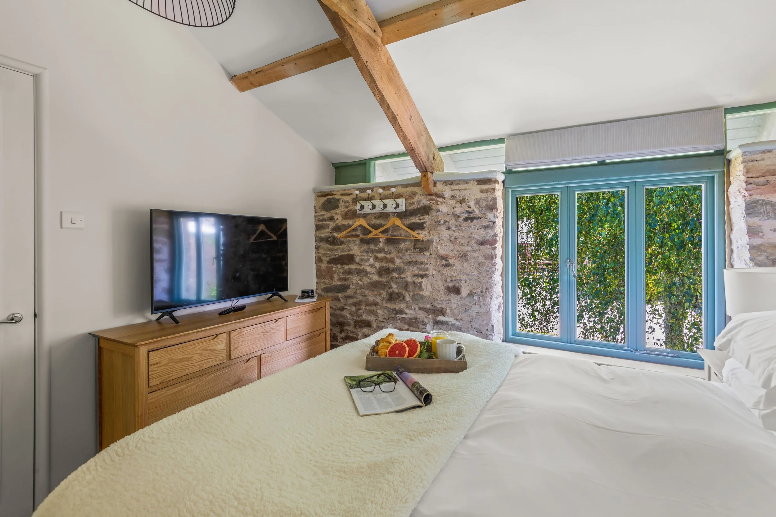 Cozy bedroom with a bed, a wooden dresser with a TV, a breakfast tray with fruit, coffee, and reading glasses, stone wall accent, large window with greenery outside, exposed wooden beam, and minimalist decor.