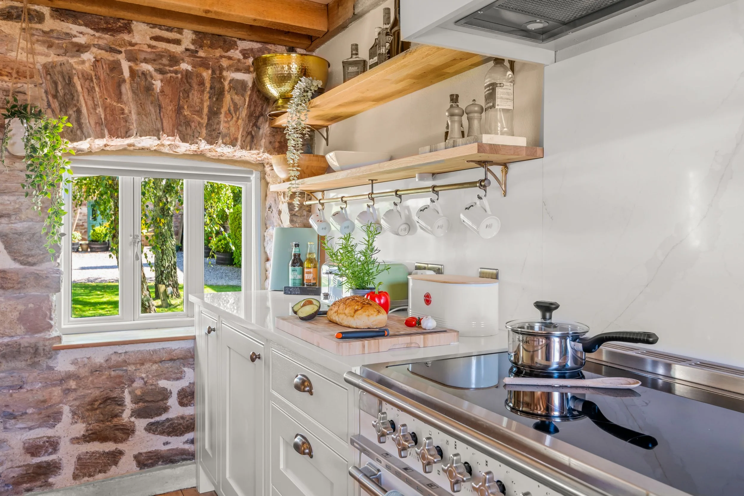 A cozy kitchen with exposed brick wall, window showing greenery outside, white cabinetry, wooden open shelves, hanging mugs, and a glass-top stove with a pot. Countertop has bread, tomato, garlic, avocado, plant, and condiments.