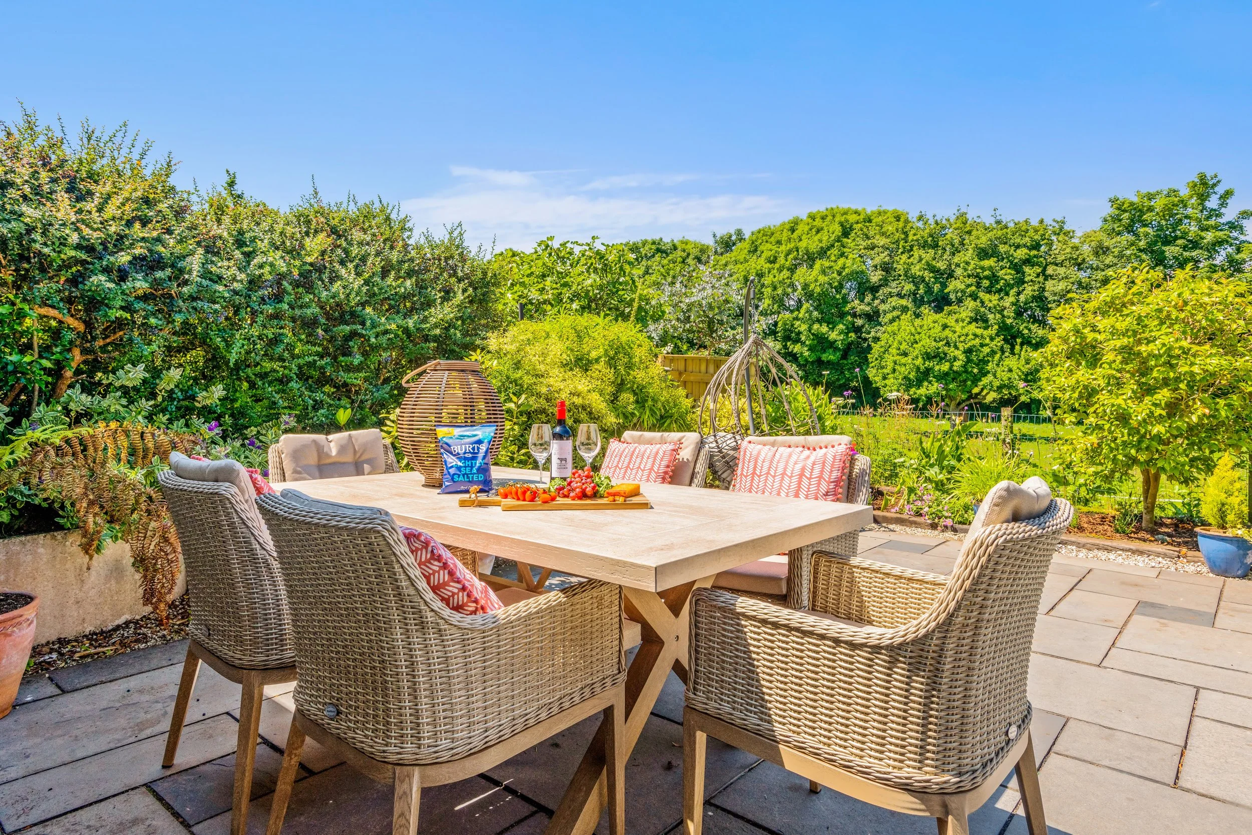 Outdoor patio with a rectangular table surrounded by six wicker chairs, some with patterned cushions, set in a lush garden with trees and shrubs under a clear blue sky.