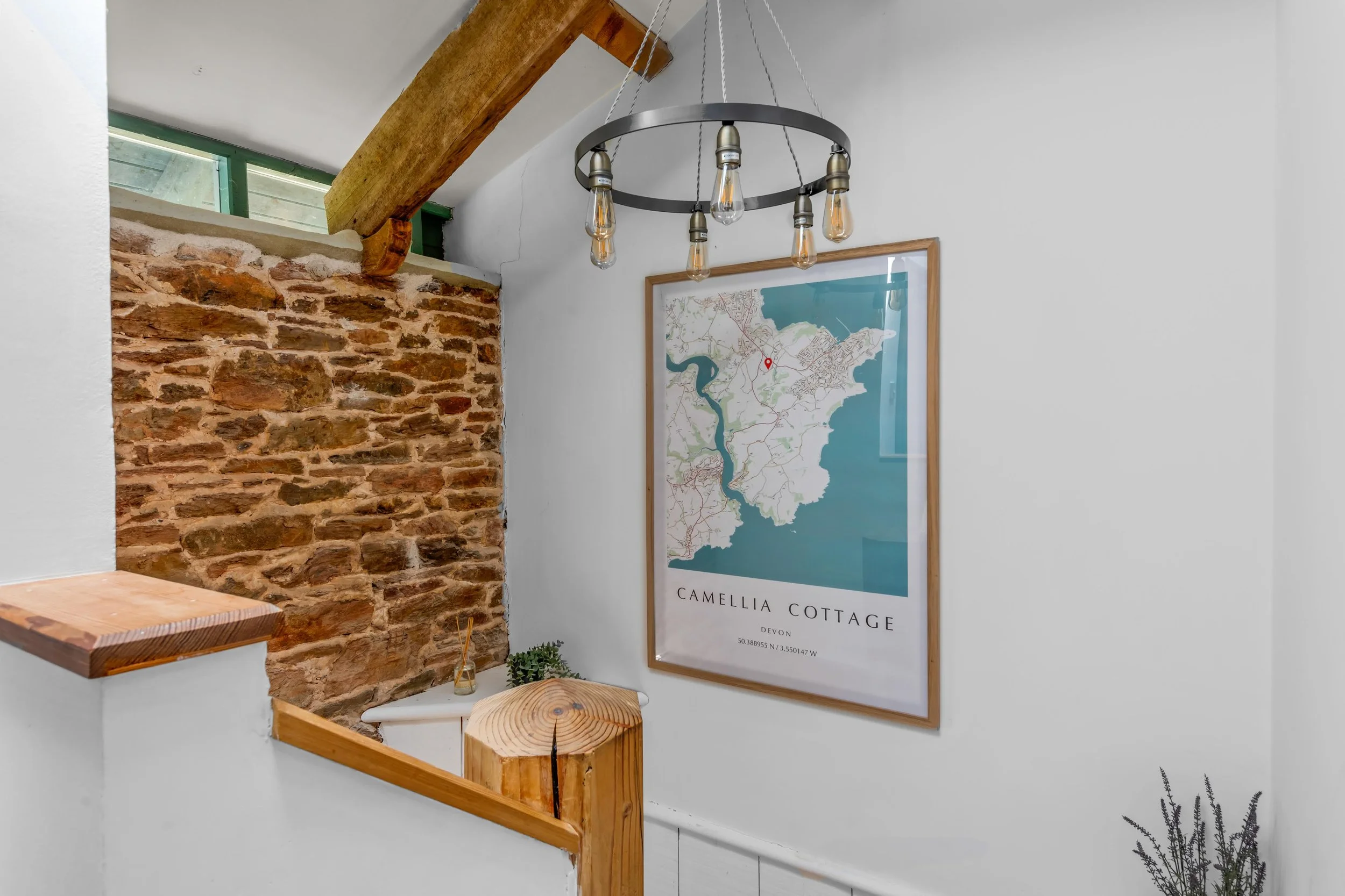 Interior view of a room corner with a white wall, exposed brick wall, wooden beam, modern chandelier with exposed bulbs, and a framed map of Camellia Cottage in Devon, UK.