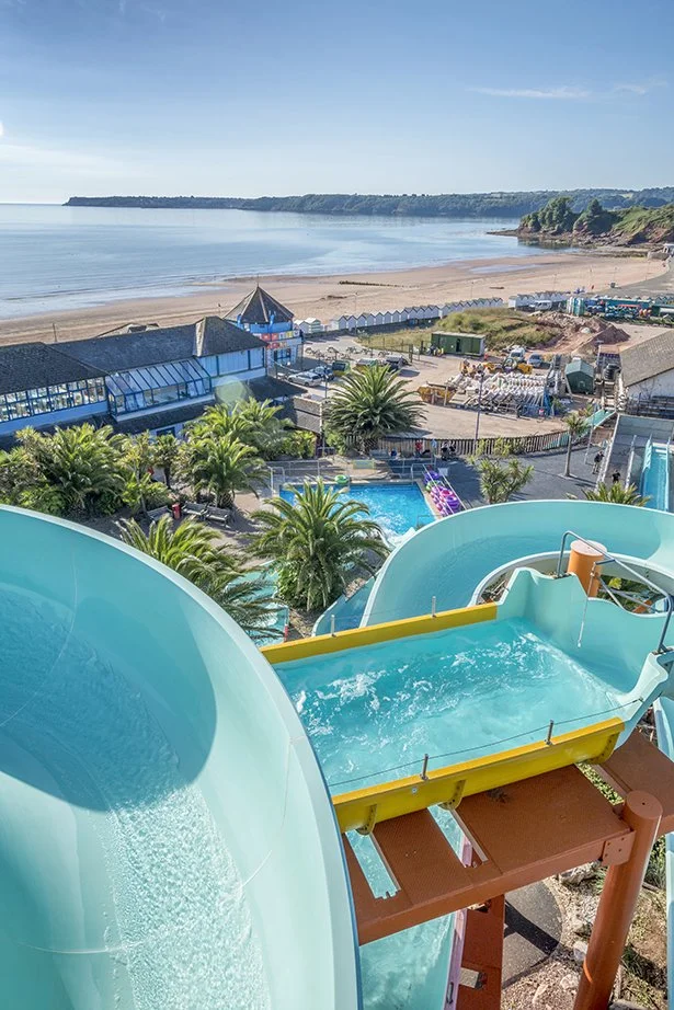 A view from a tall water slide looking down at a water park with a pool, palm trees, and a beachside ocean in the background.