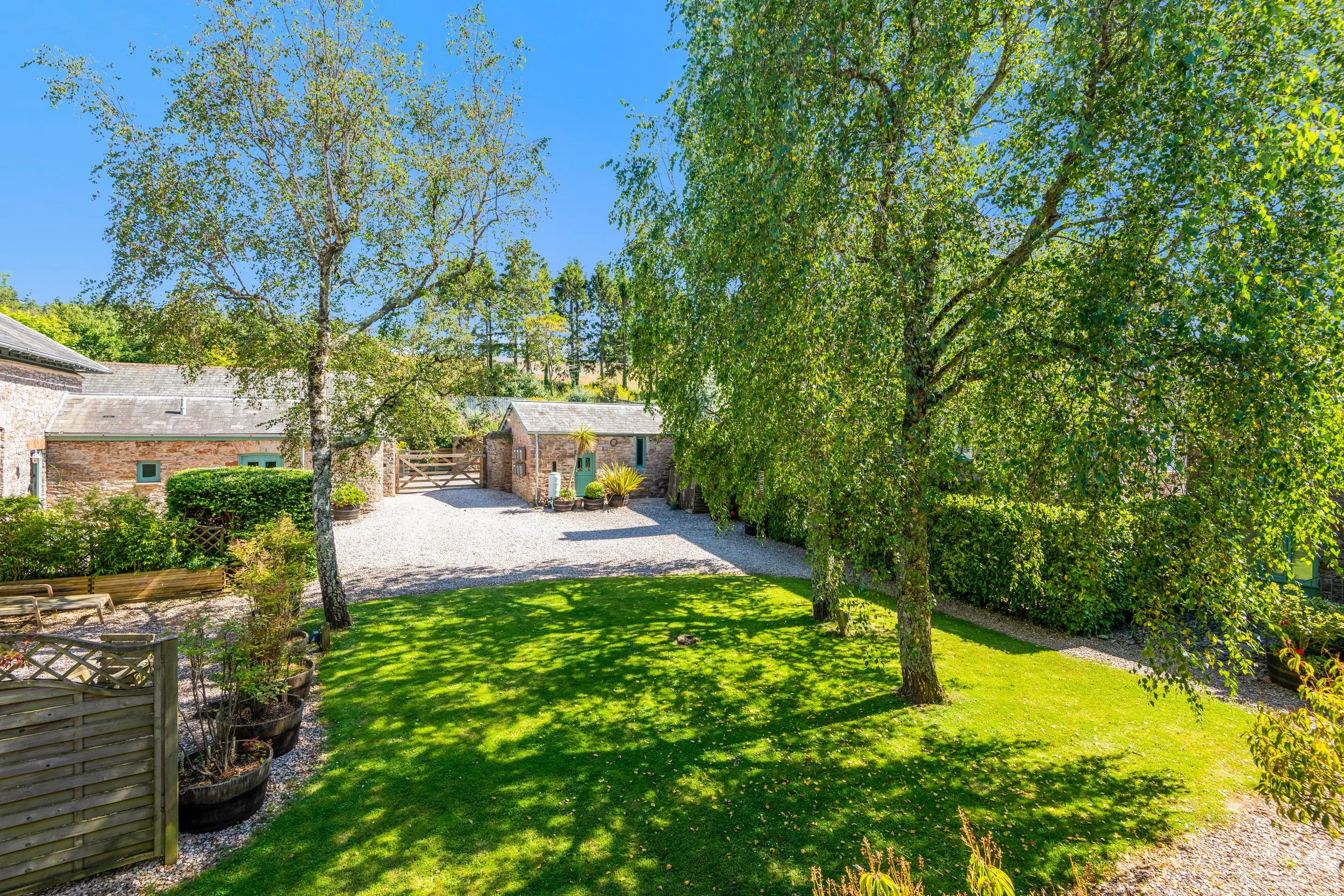 A peaceful garden scene with lush green grass, tall leafy trees casting shadows, and a gravel pathway leading to a stone building with a gate under a bright blue sky.