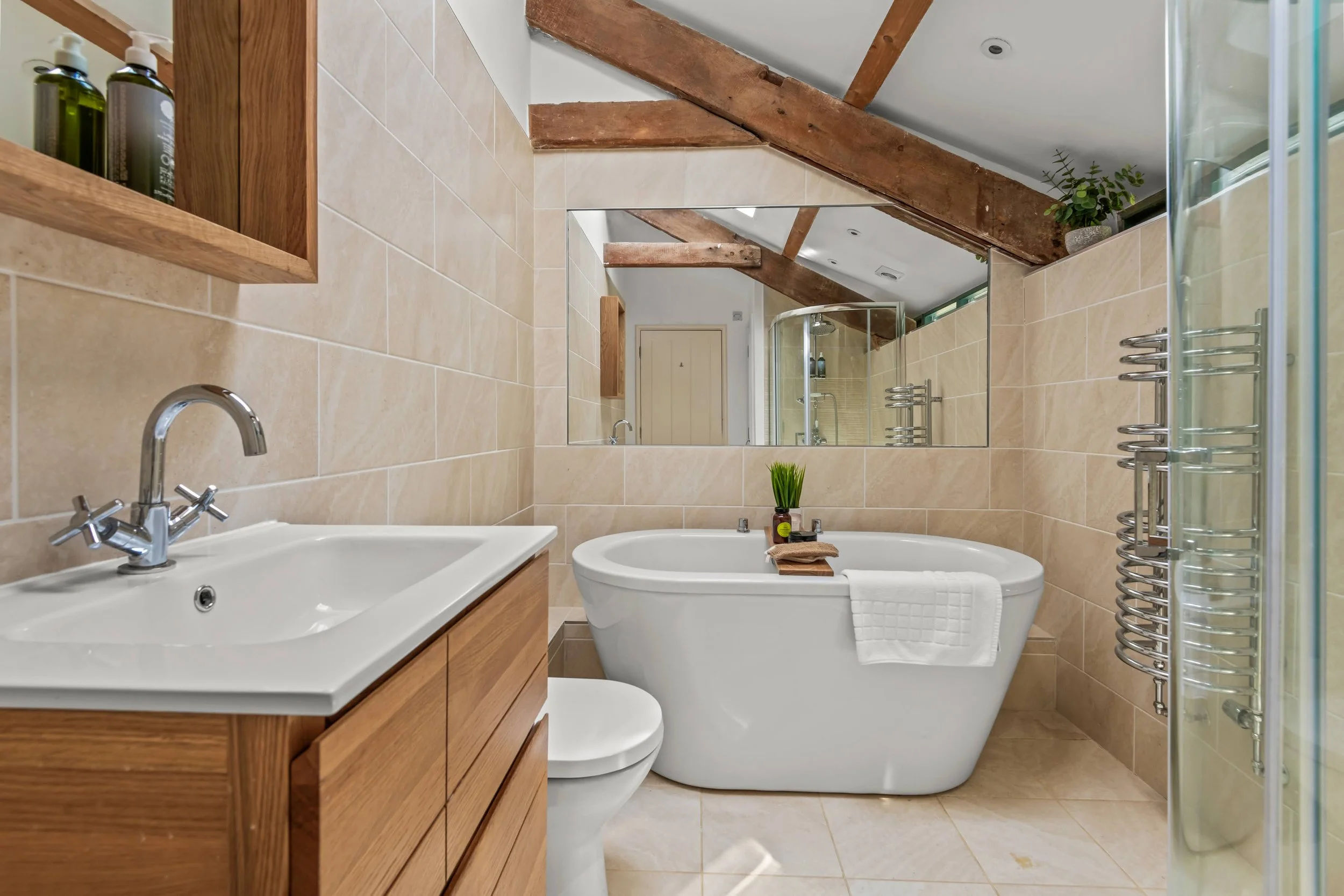 A modern bathroom with a wooden vanity, a white bathtub with a towel, a mirror, and a glass shower enclosure, featuring exposed wooden beams on the ceiling and a beige tiled wall.