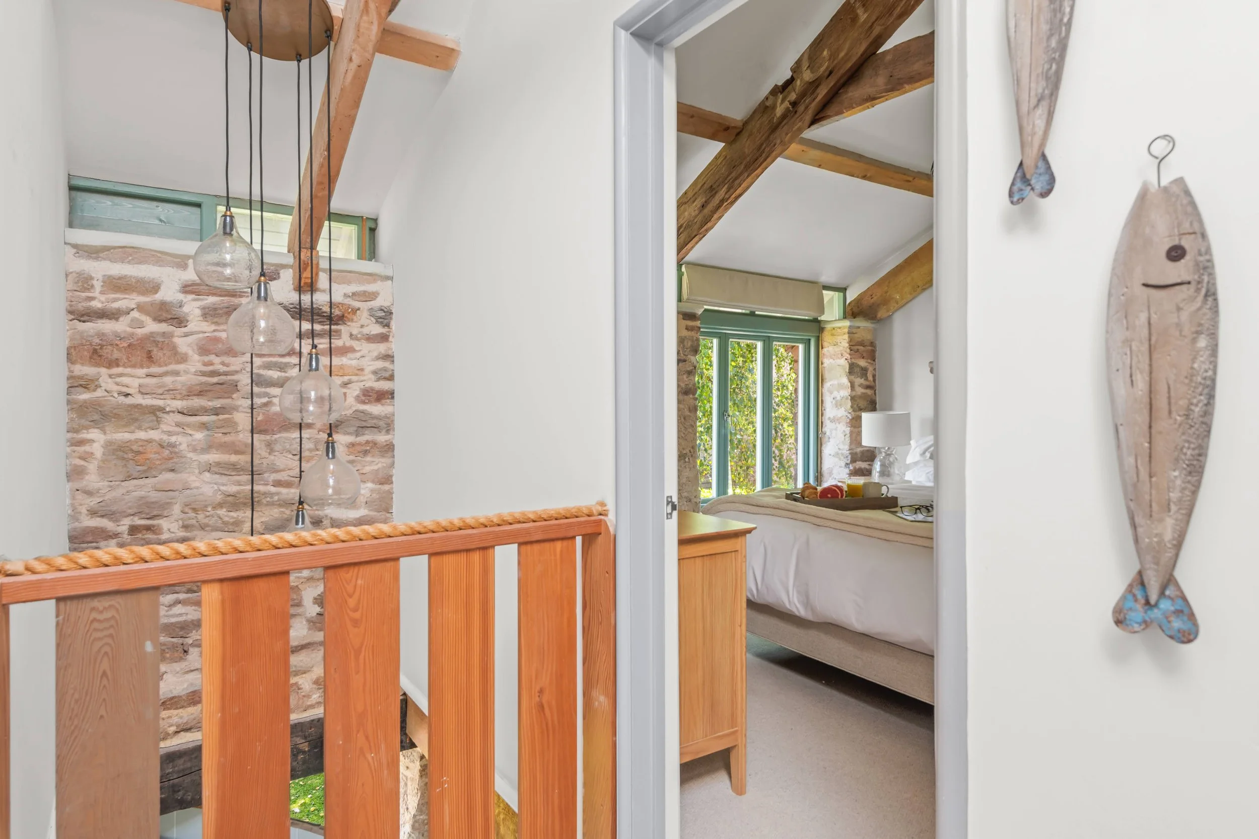 View of a bedroom through an open doorway, featuring wood beams on the ceiling, a large window with greenery outside, and a bed with white bedding and a tray of breakfast on it. A small wooden nightstand with a white lamp is beside the bed. To the le