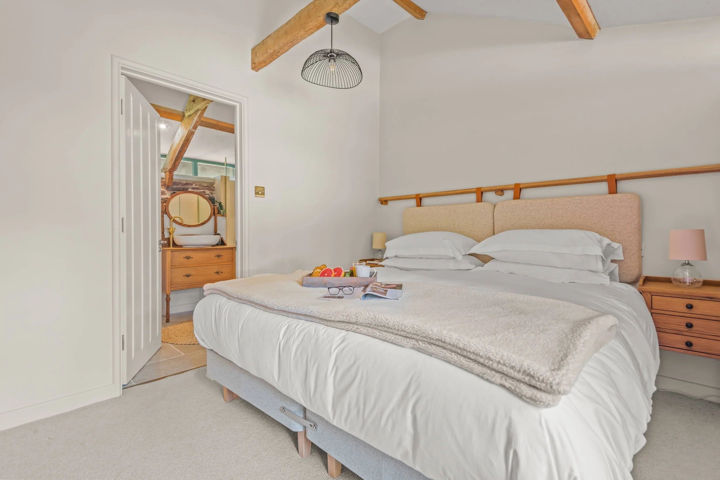 A cozy bedroom with a made bed, pillows, and a tray of fruit and a magazine. Wooden beams and a pendant light add charm. An open door shows a bathroom with a vanity and mirror.