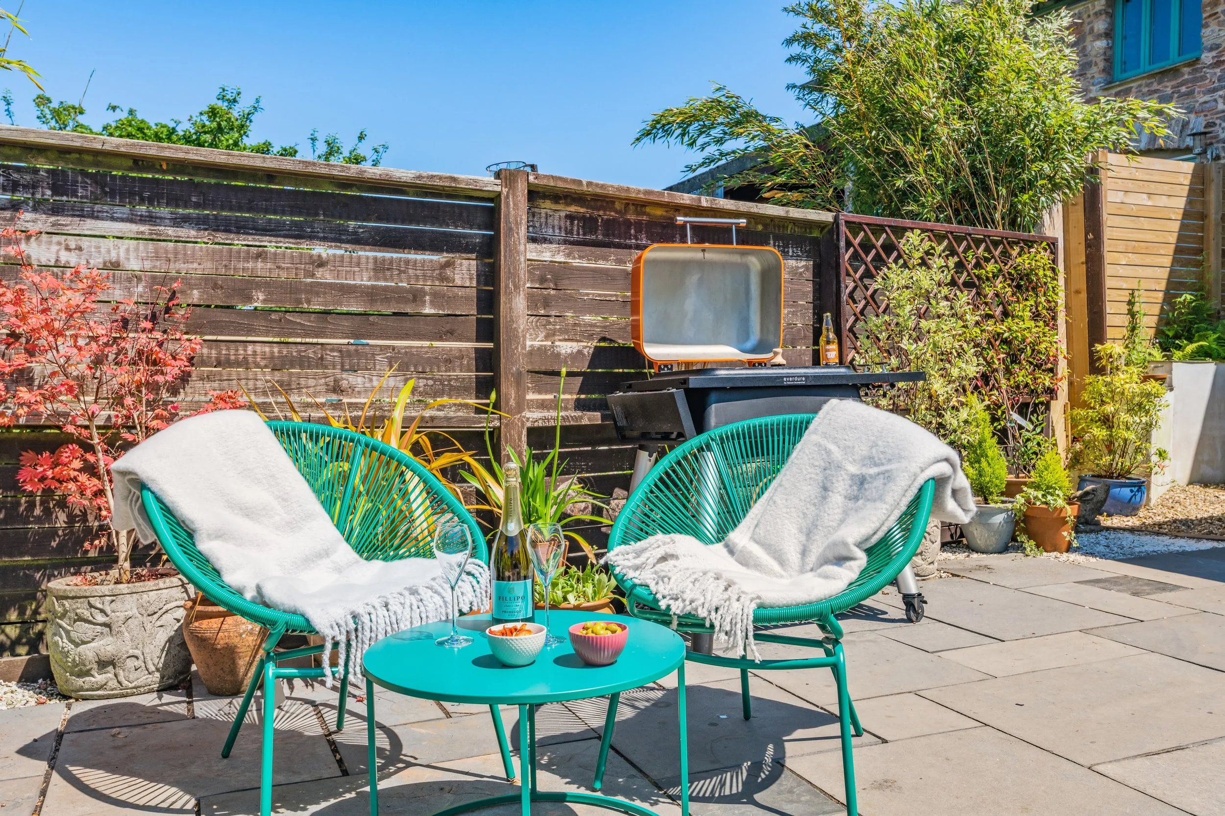 A cozy outdoor patio area with two turquoise lounge chairs with white towels, a small turquoise table with snacks and drinks, a barbecue grill, and potted plants against a wooden fence on a sunny day.