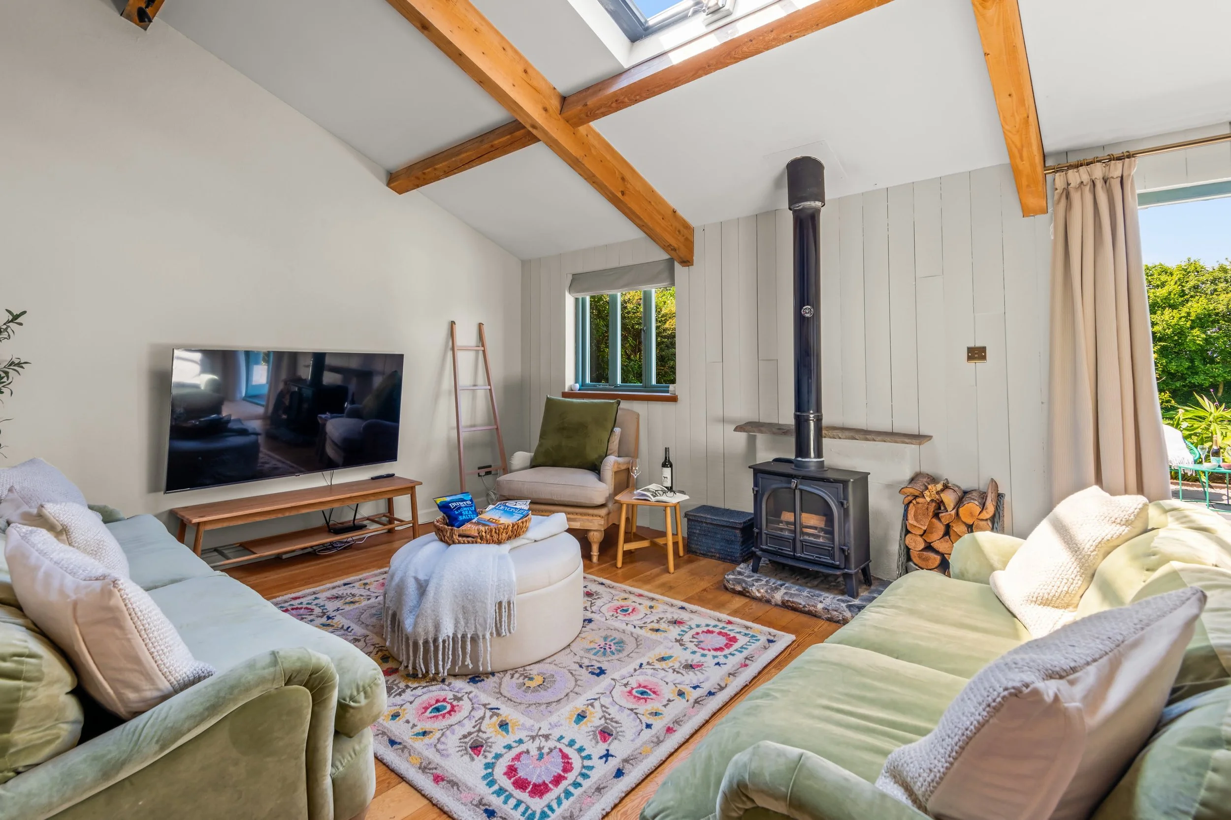Living room with green sofas, a flat-screen TV, a patterned rug, a wood-burning stove, and large windows with views of trees.