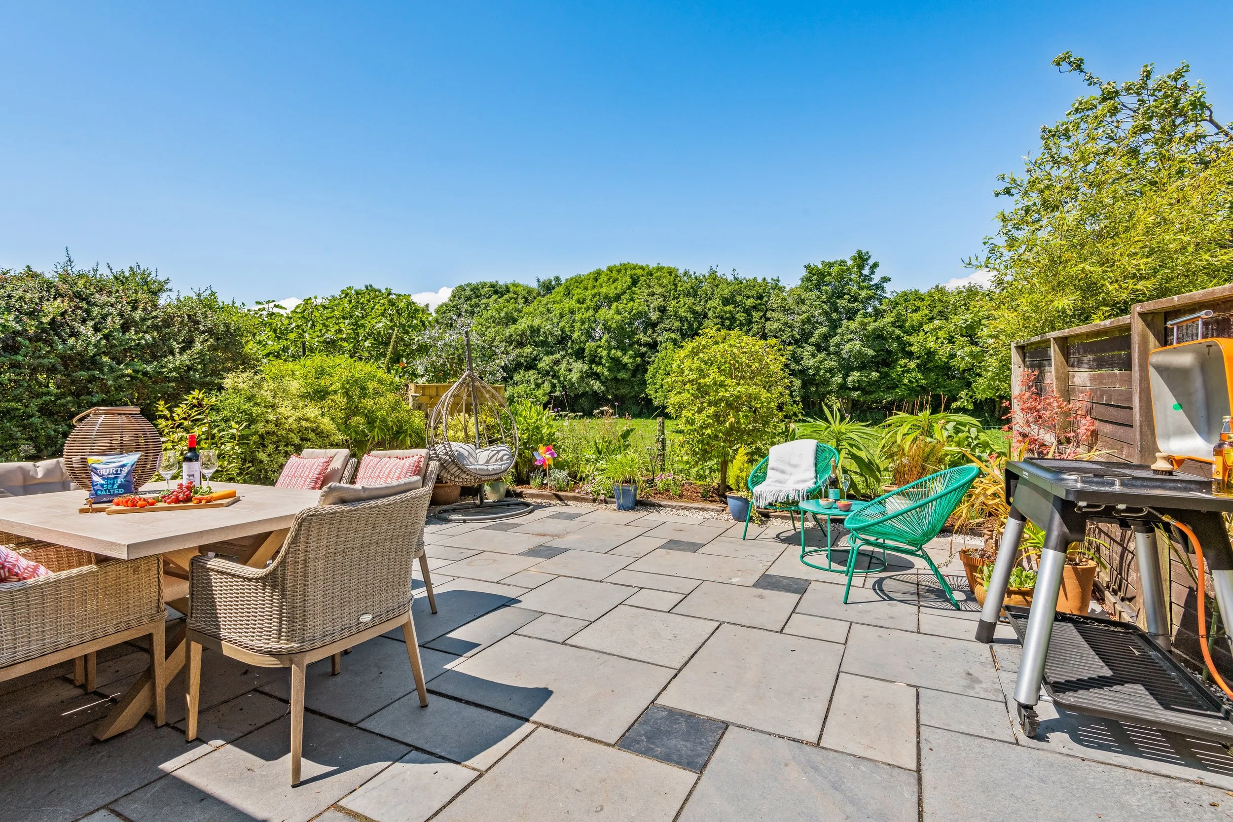 A spacious outdoor patio with a dining table, wicker chairs, a hammock swing, potted plants, a grill, and garden greenery under a clear blue sky.