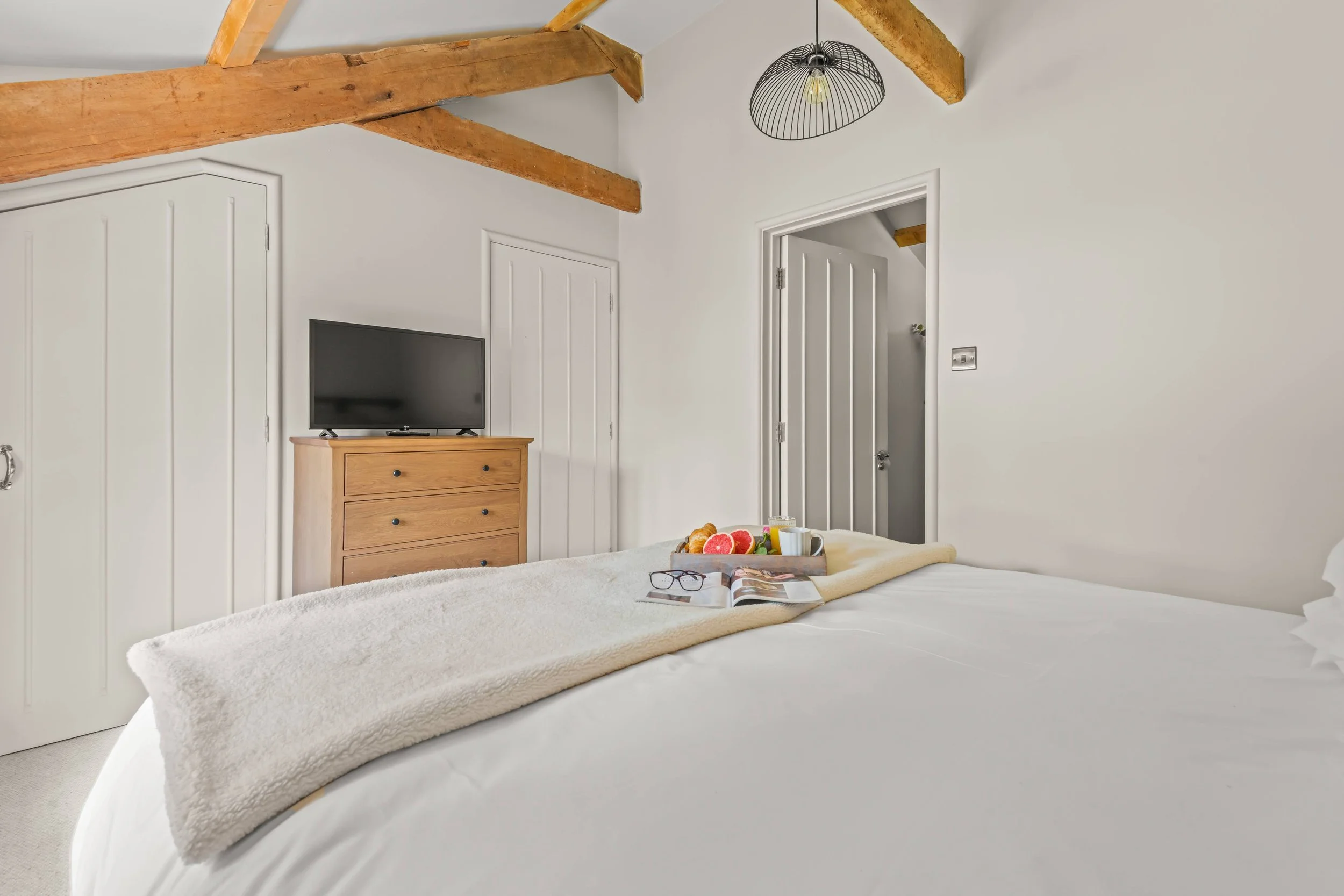 A cozy bedroom with white walls and wooden ceiling beams. A bed with white bedding has a beige blanket and a breakfast tray with fruit, coffee, and reading glasses. There is a wooden dresser with a TV on top and two closed white closet doors. An open