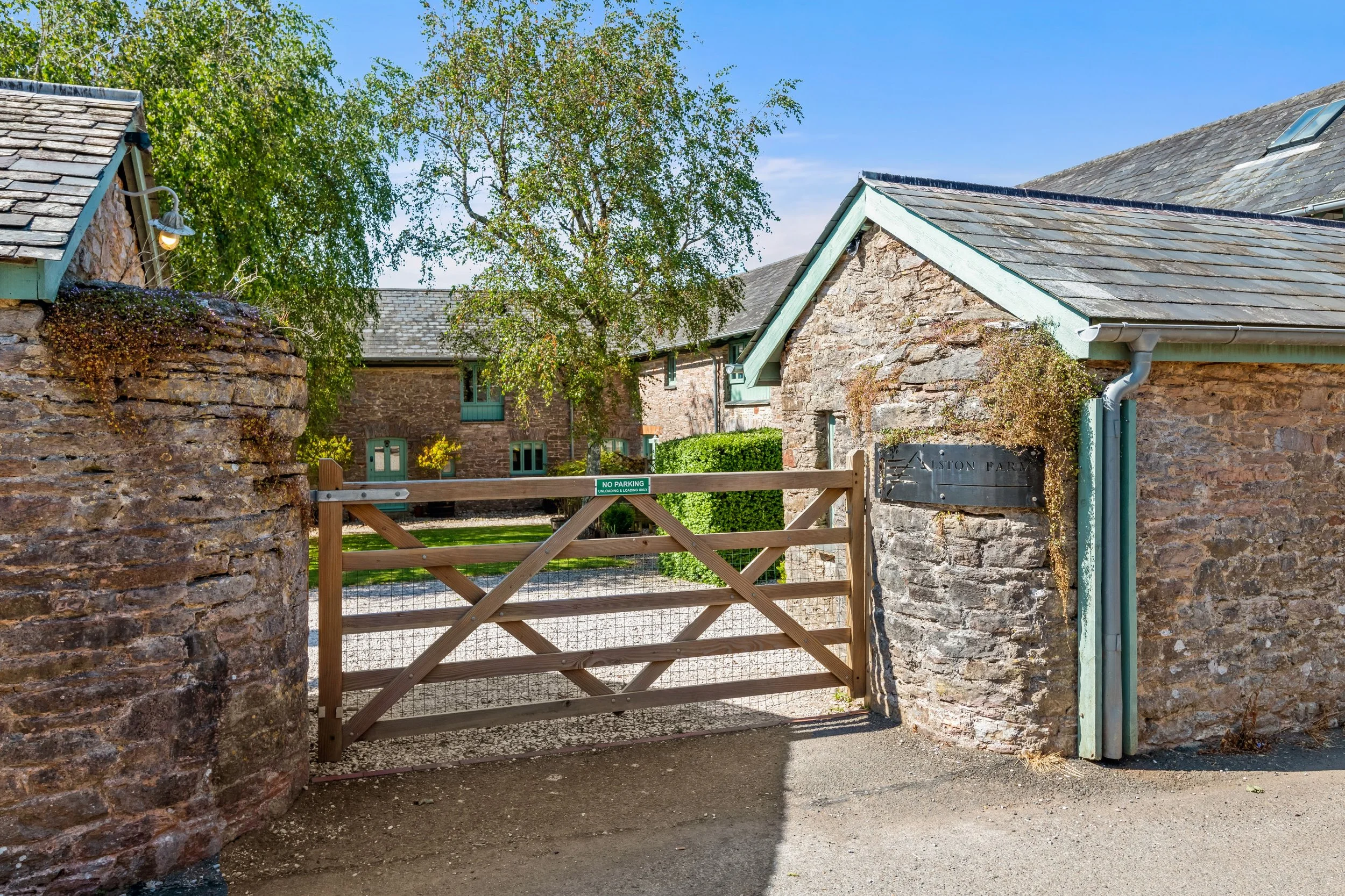 Stone entrance gate with a wooden fence, green windows, and trees in the background at a farm or rural property.