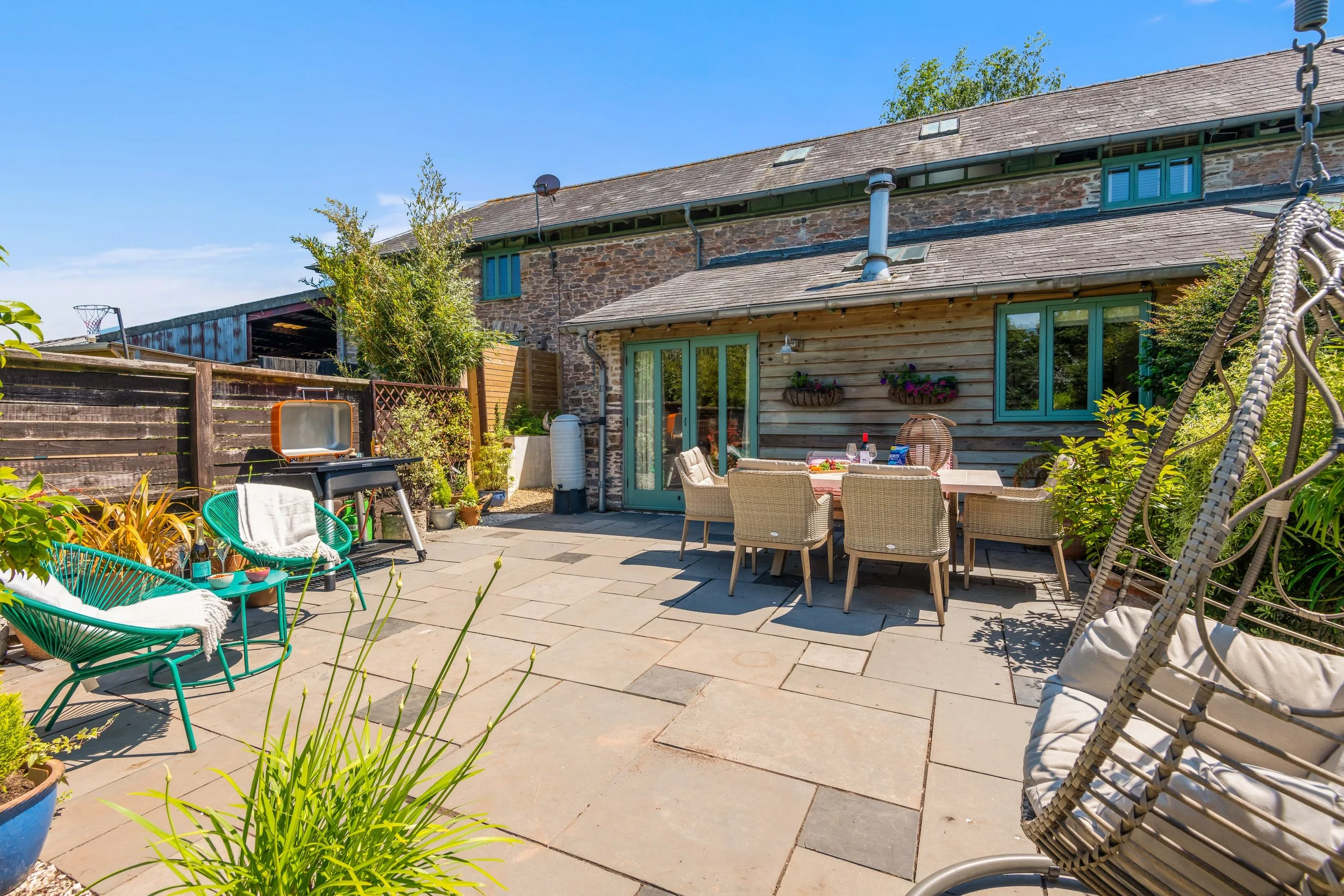 Outdoor patio with dining table, chairs, potted plants, swing chair, and a barbecue grill against a house with stone and wood siding under a blue sky.