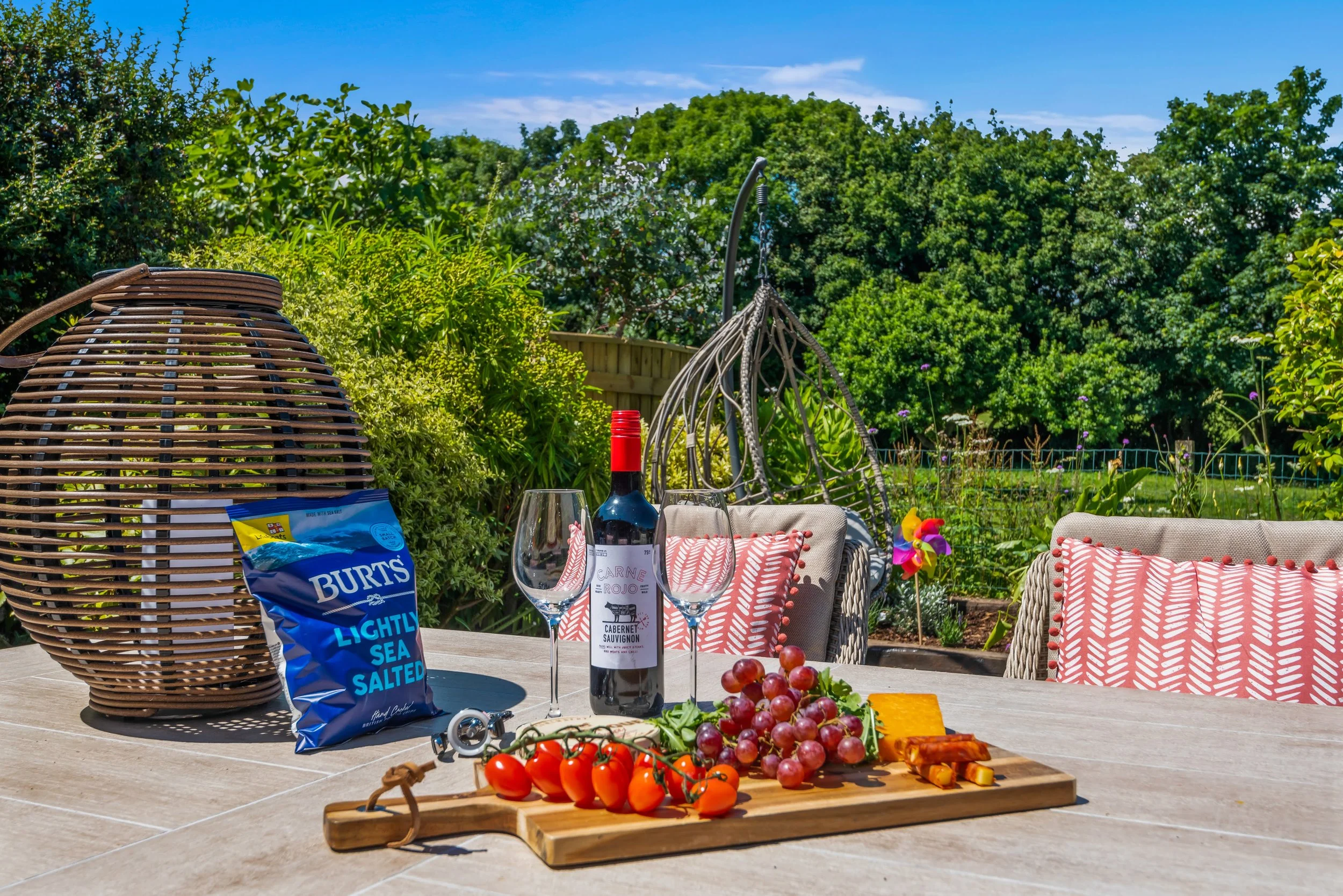An outdoor patio table setup with a bottle of red wine, two wine glasses, a cutting board with cherry tomatoes, grapes, cheese, and crackers. In the background, there are wicker chairs with red and white patterned cushions, a hanging egg-shaped chair