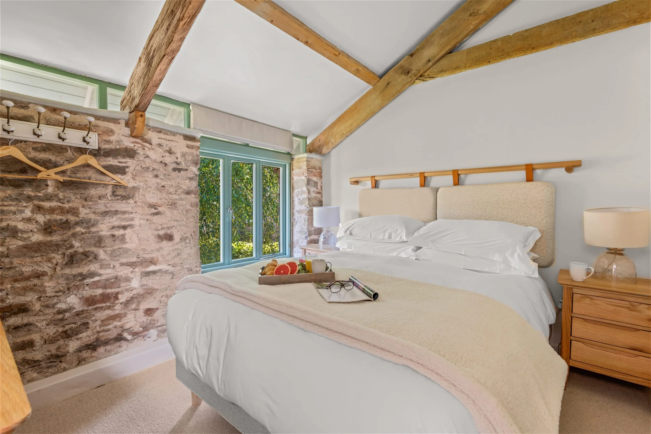 A cozy bedroom with a large bed featuring a beige headboard, a tray of breakfast items, and exposed wooden beams on the ceiling. There are two nightstands with lamps and a window with greenery outside.