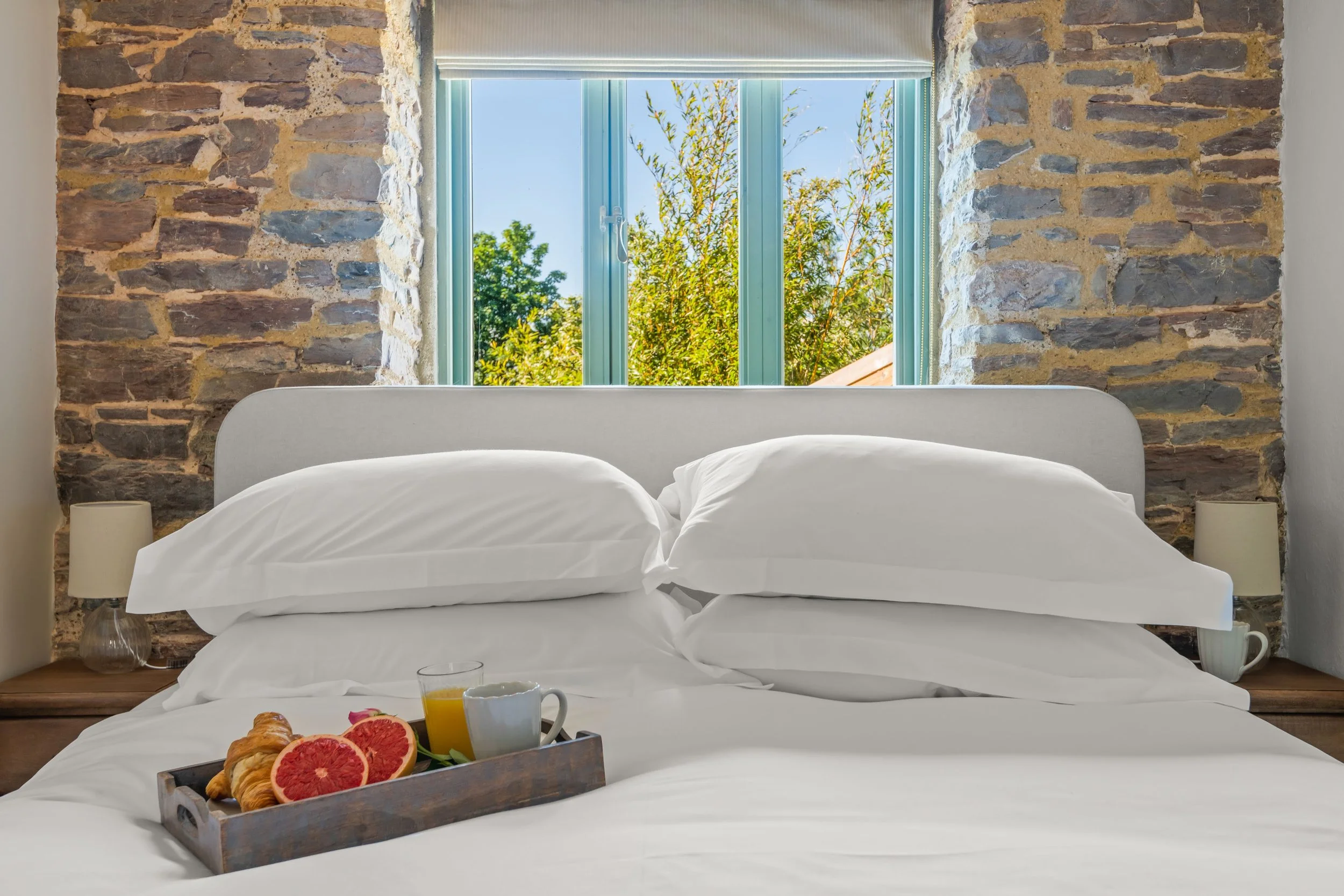 A neatly made bed with white pillows and bedding, featuring a breakfast tray with grapefruit slices, a croissant, an orange juice glass, and a coffee mug, in a room with a stone wall and a window showing green trees outside.