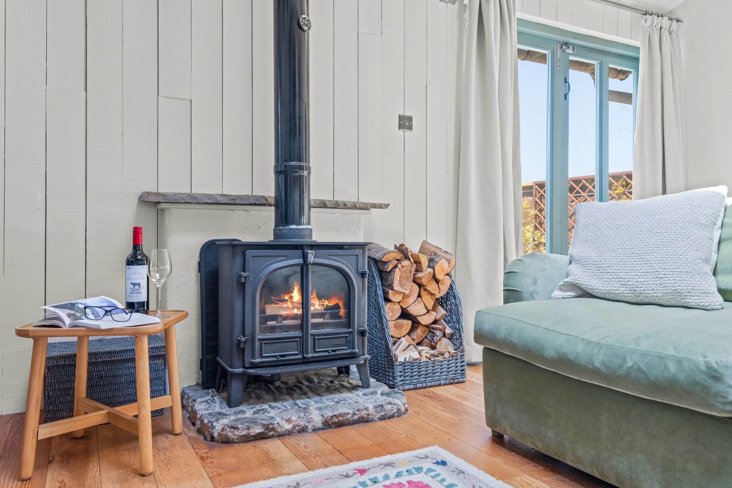 Living room with a wood-burning stove, a basket of firewood, a small wooden side table with a bottle of wine, a glass, and glasses, a green sofa with pillows, and a large window with white curtains showing a sunny outdoor scene.