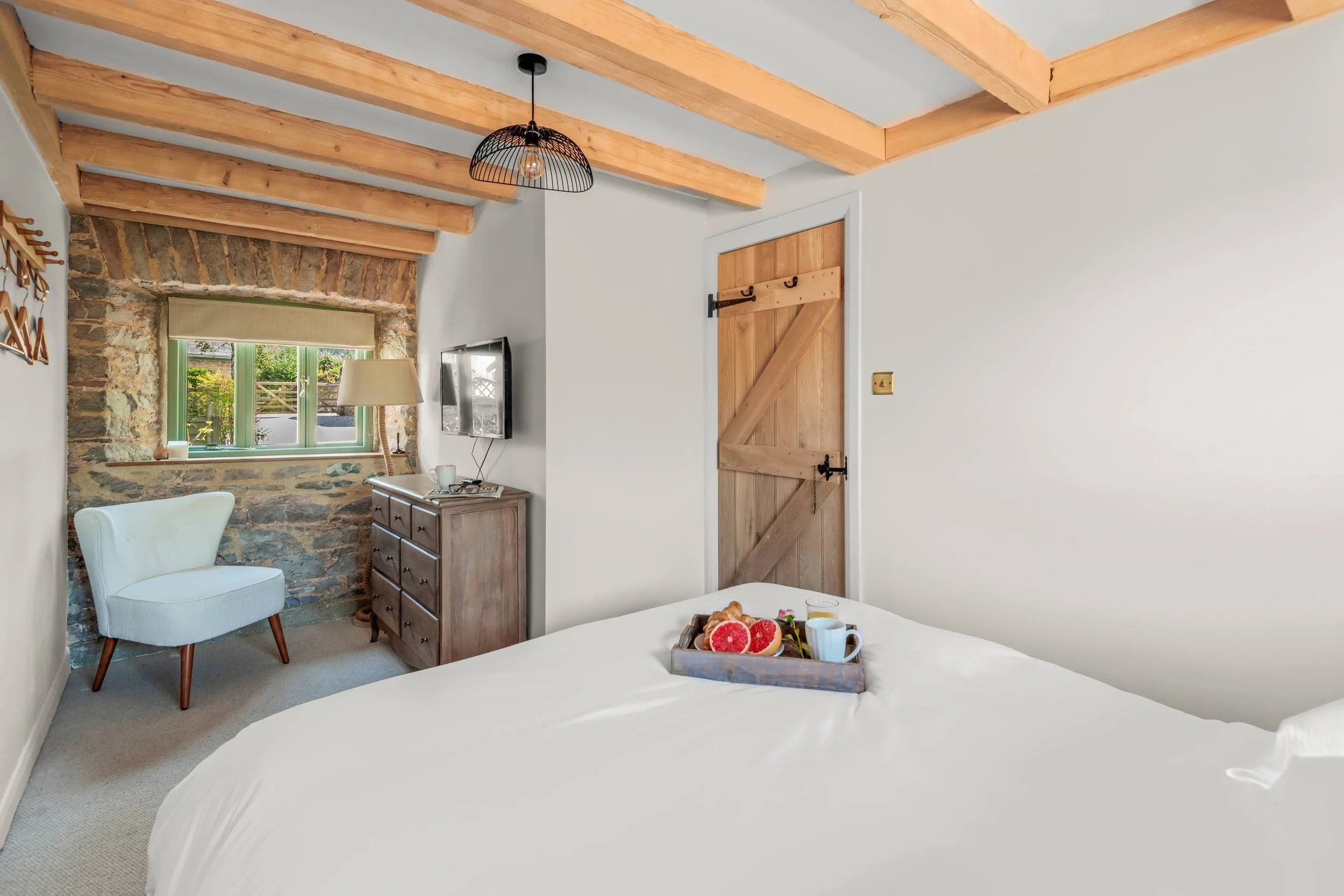 Cozy bedroom with white walls, a wooden ceiling with exposed beams, a wooden door, a small dresser with a lamp, a wall-mounted TV, a chair near a stone wall window, and a breakfast tray on the bed with grapefruit, coffee, and a glass of water.