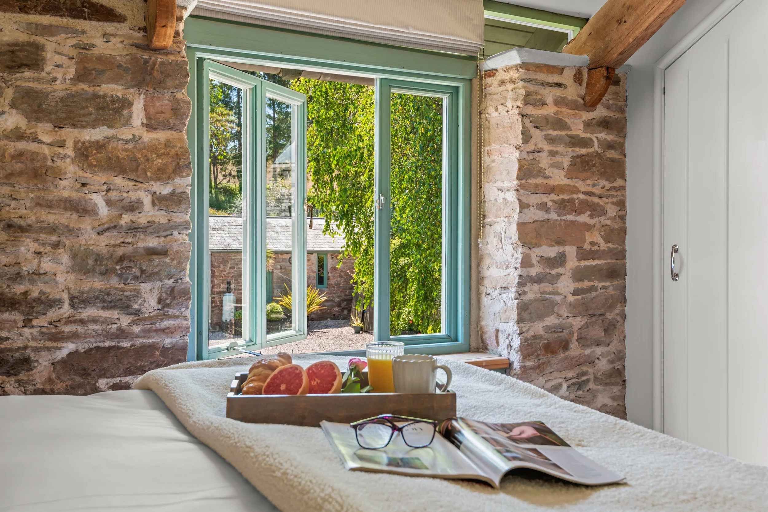A cozy bedroom with a ledge table in front of a window showing a garden outside. The table has a tray with grapefruit, a glass of orange juice, a white mug, and a small vase with a pink flower. On the bed, there is an open magazine and a pair of blac