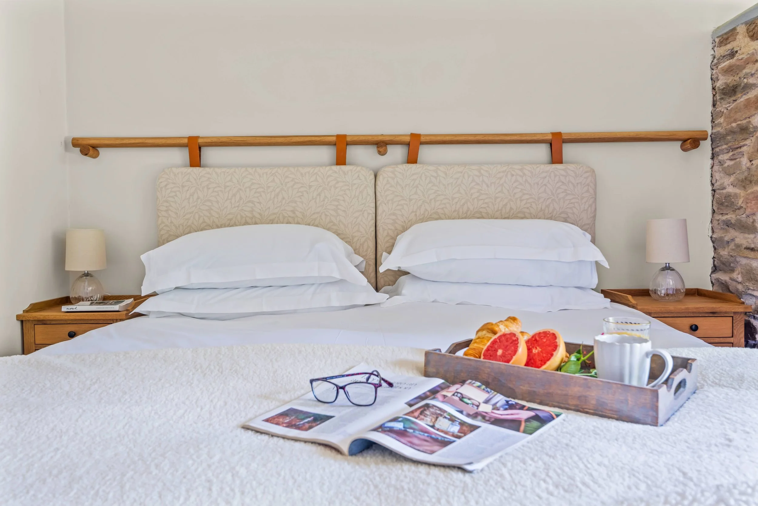 Hotel bed with white bedding, magazine, glasses, a tray with grapefruit, croissants, a cup, and a glass of water, two bedside tables with lamps, and a stone accent wall.