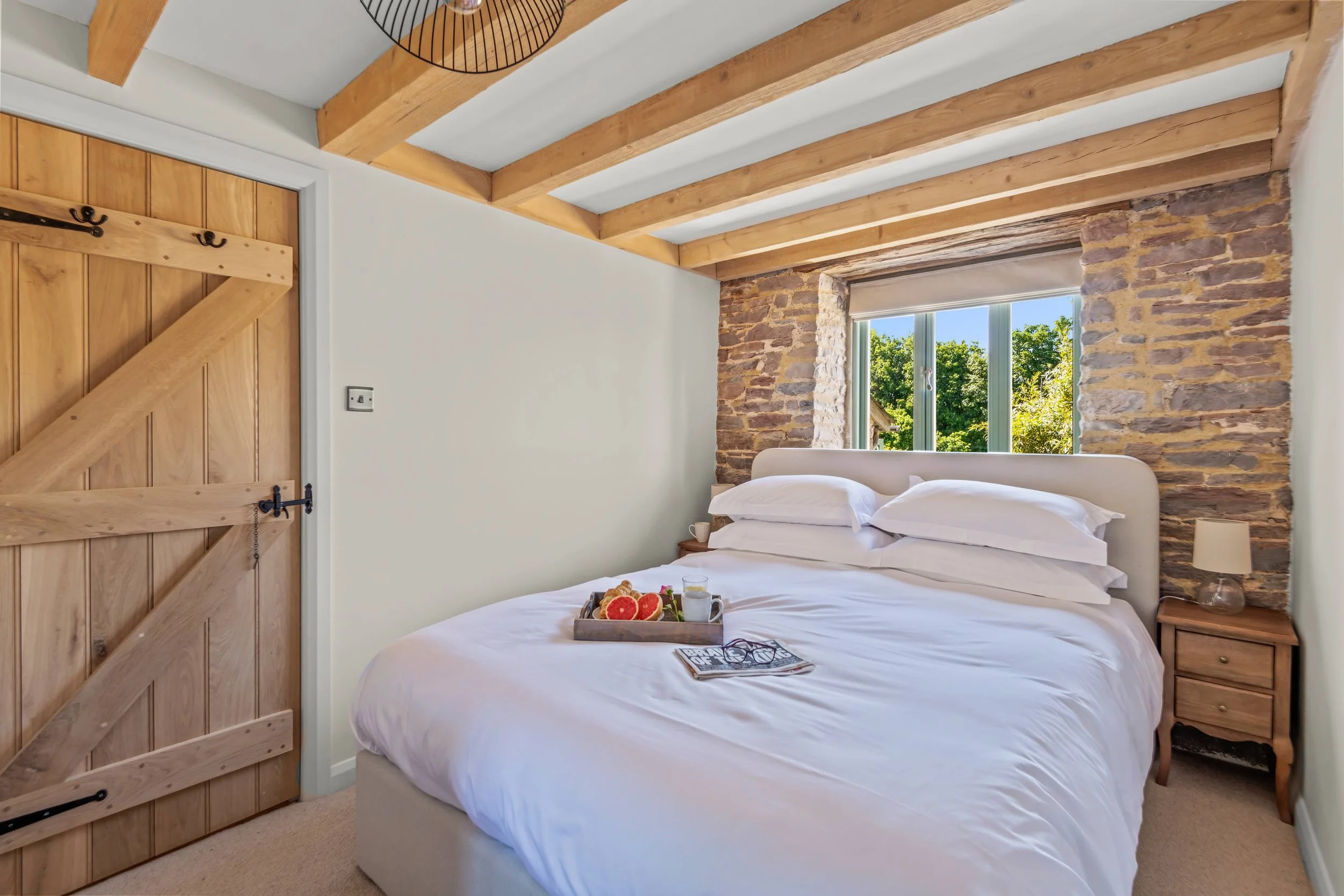 Bedroom with a white bed, a tray of breakfast items, and a window showing green trees outside, with exposed brick and wooden beams ceiling.