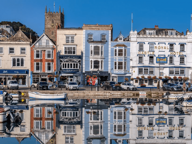 Colorful buildings along a waterfront with boats and parked cars, including a hotel named 'Royal Castle Hotel' and a restaurant, with their reflections visible in the water.