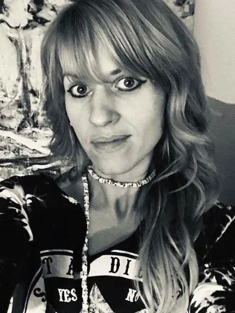 A woman with wavy hair and bangs, wearing a choker necklace, a patterned shirt, and looking into the camera in a selfie.