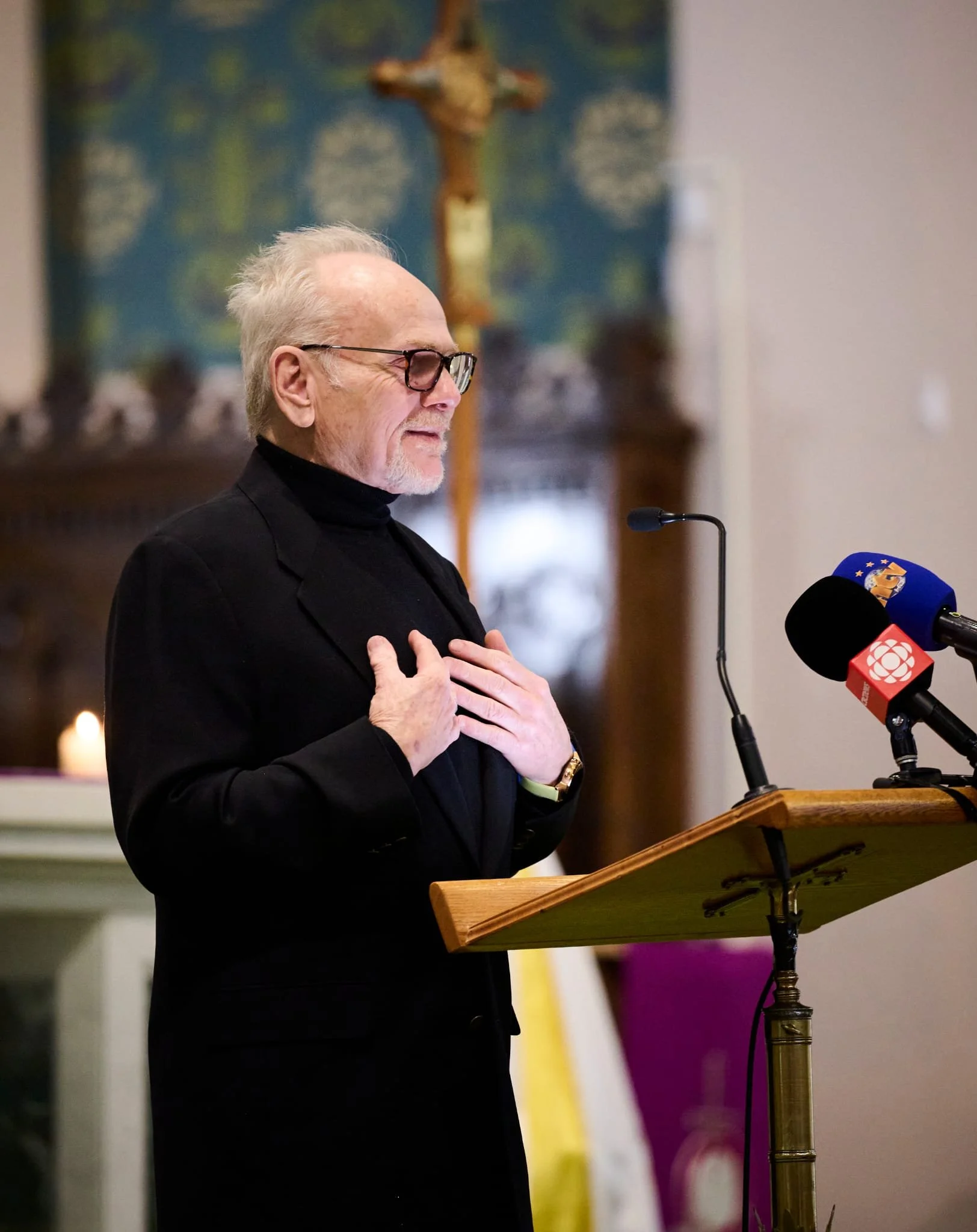 An elderly man with glasses, dressed in a black suit and turtleneck, is standing at a wooden pulpit with microphones, holding his chest with one hand while the other is raised. Behind him, there is a large Christian cross and religious artwork in a church setting.