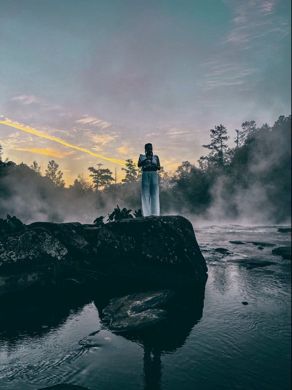 Person standing on a large rock in the middle of a river or stream, surrounded by mist and trees at sunset or sunrise.