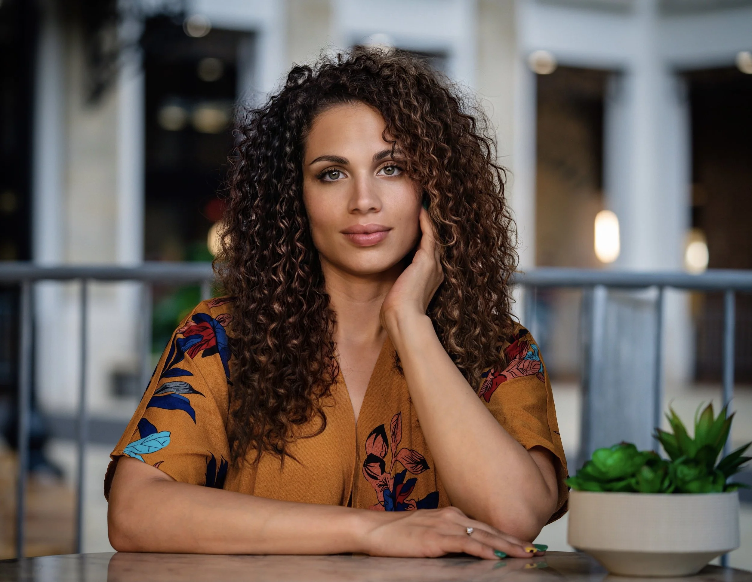 A woman with curly brown hair and light skin poses with her hand near her face, sitting at a table with a green potted plant, in a well-lit indoor setting.