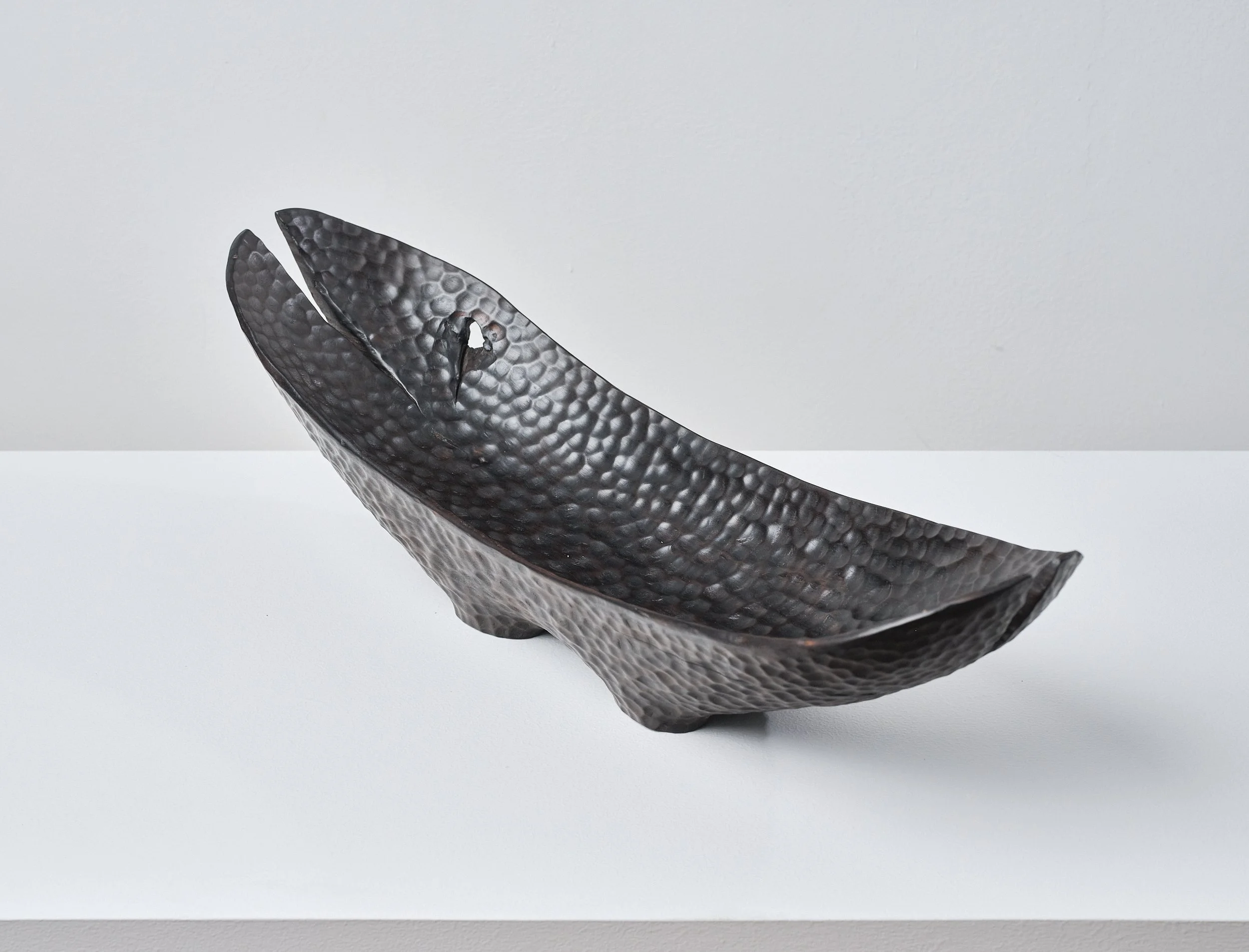 Norwegian Maple Long Bowl with dimpled polished surface that resemble black hammered metal with splits from the timber on white surface against plain white wall.
