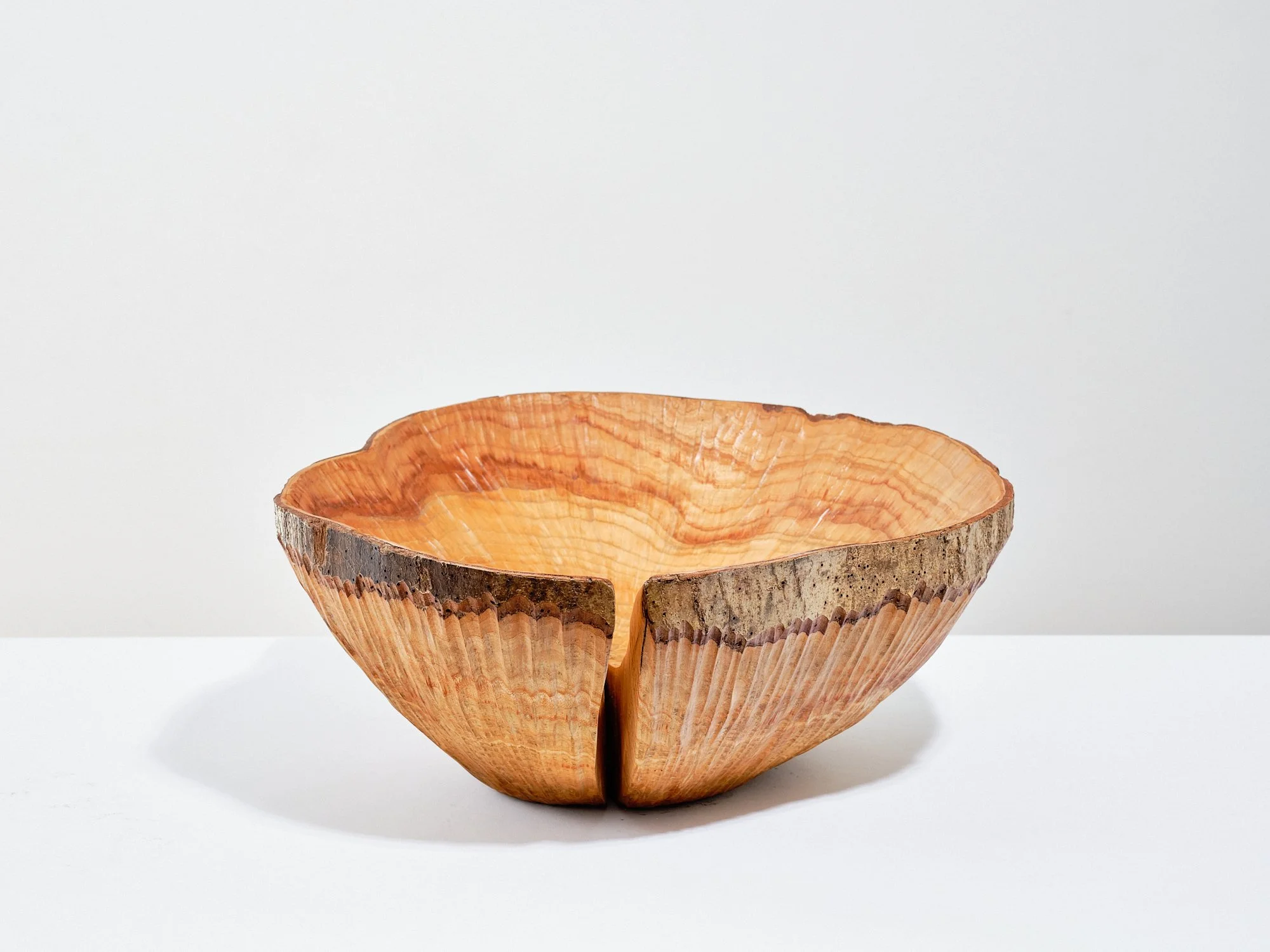 A sculptural wooden bowl showing strong radial grain with large split, made from ash with a ring of bark at the top and smooth textured interior, placed on white surface against white background