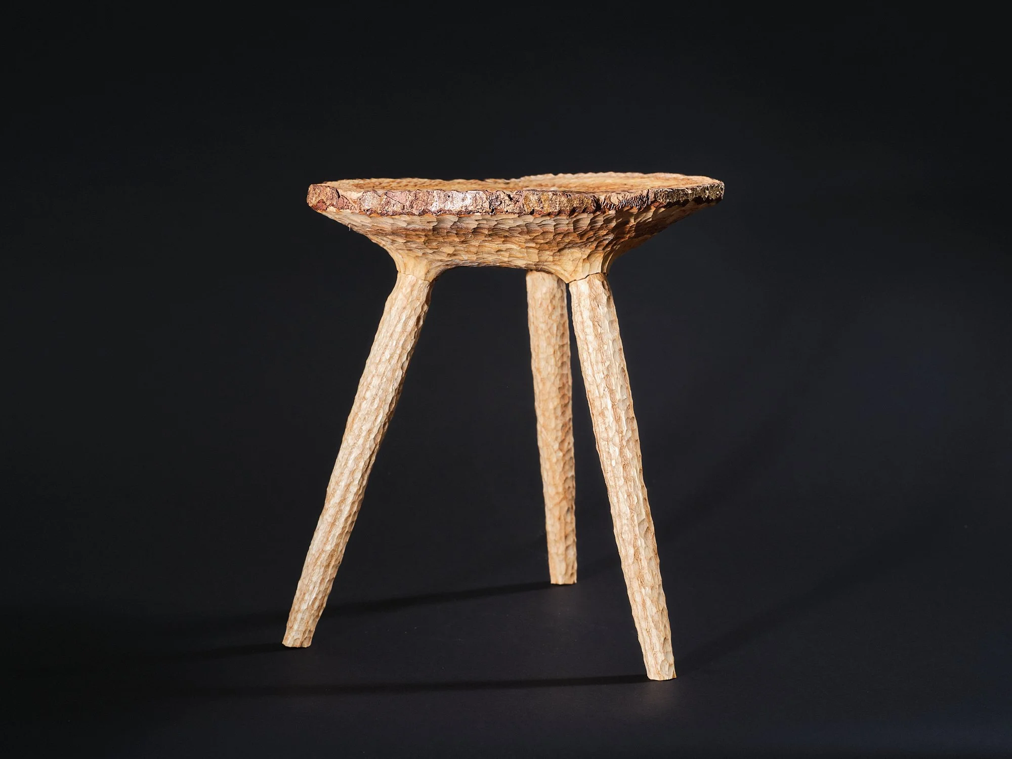 A small free carved, contemporary crafted, wooden milking stool made from a disc of ash with edge grain and three angled ash legs, oil finished, set against a black background.