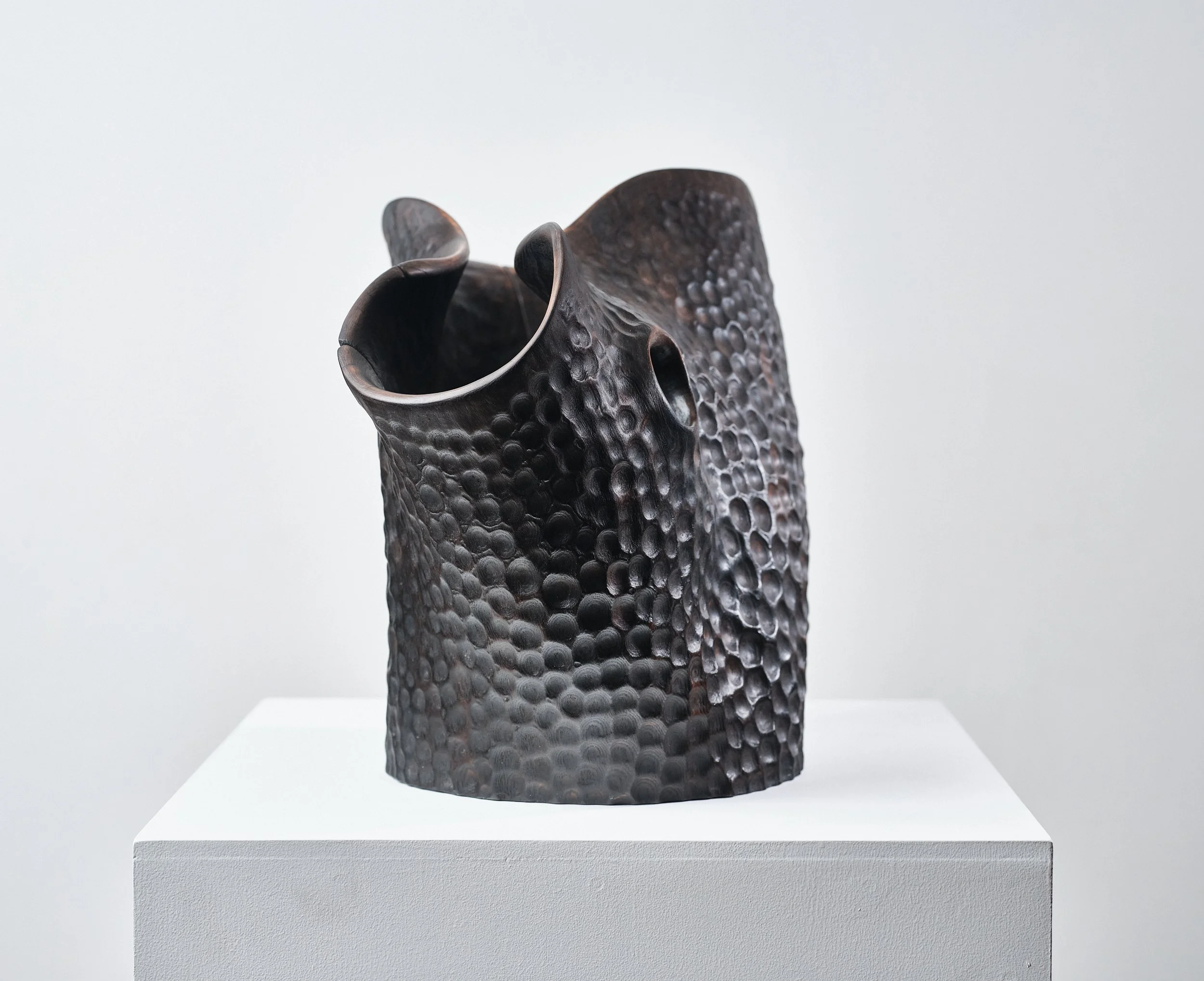 A black textured carved wood functional art piece with an irregular, ruffled opening, displayed on a white pedestal against a plain background.