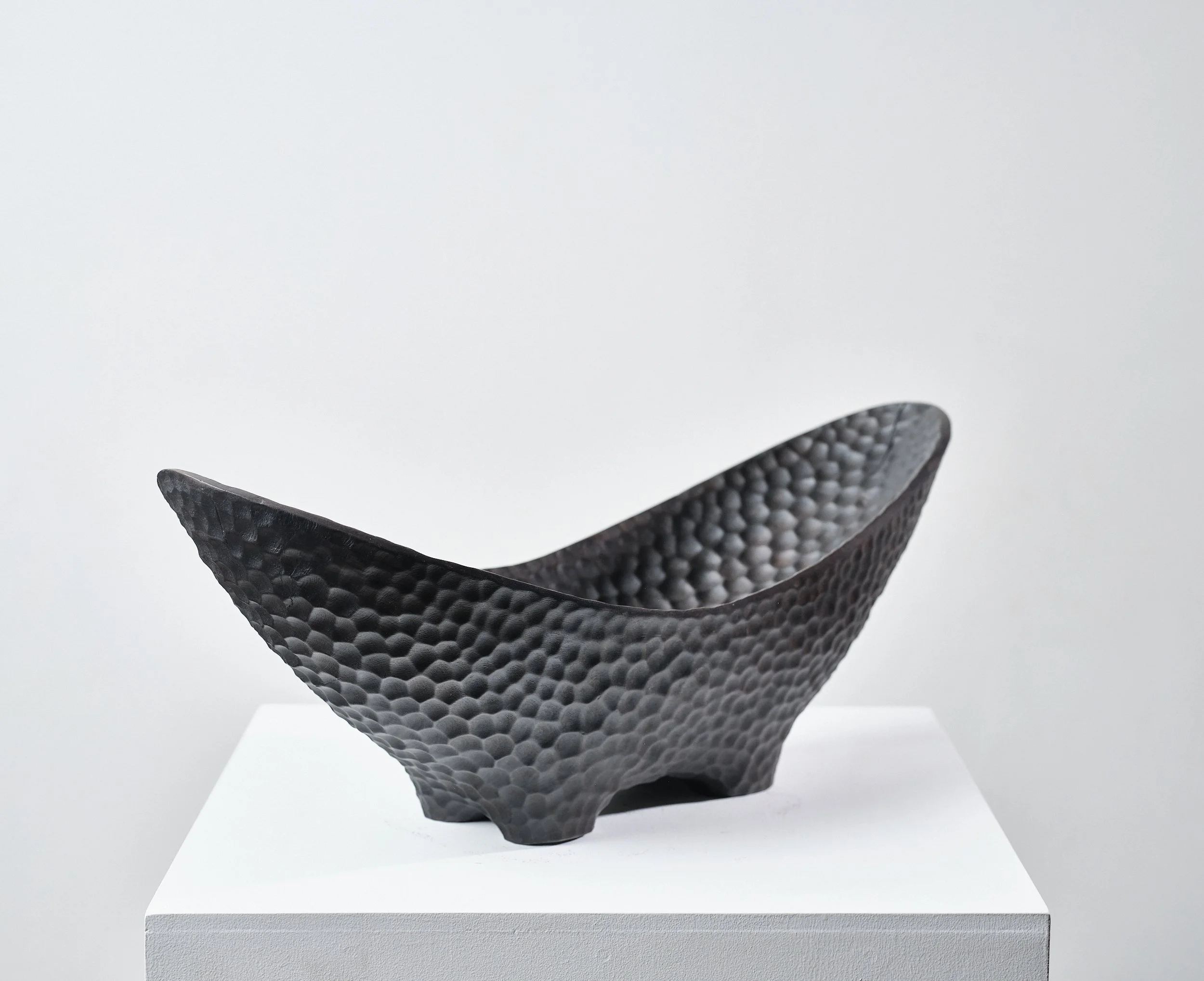 Black textured bowl made from scorched and waxed London Plane timber with an organic shape, displayed on a white pedestal in a gallery setting.