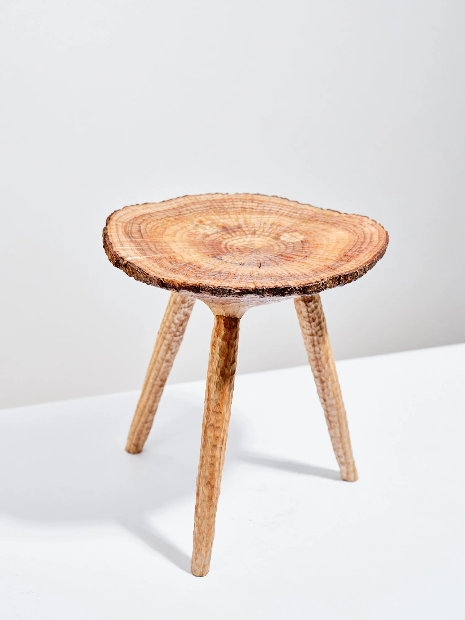 A small free carved, contemporary crafted, wooden milking stool made from a disc of ash with edge grain and three angled ash legs, oil finished, set against a white background.