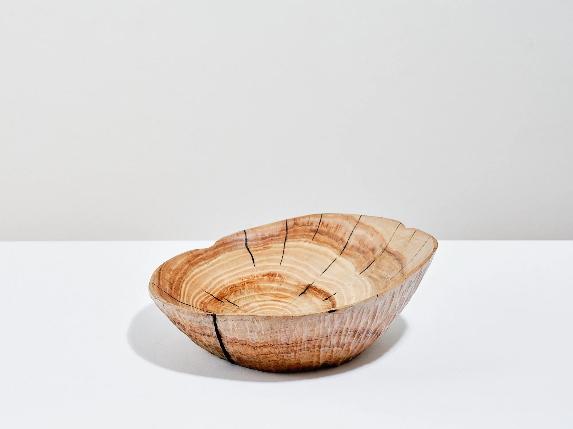 A seasoned ash bowl in a natural waxed and polished wood colour with cracks and splits repaired Kintsugi-style using black pine resin glue, providing visual contrast to the rest of. placed on a white surface against a plain white background.