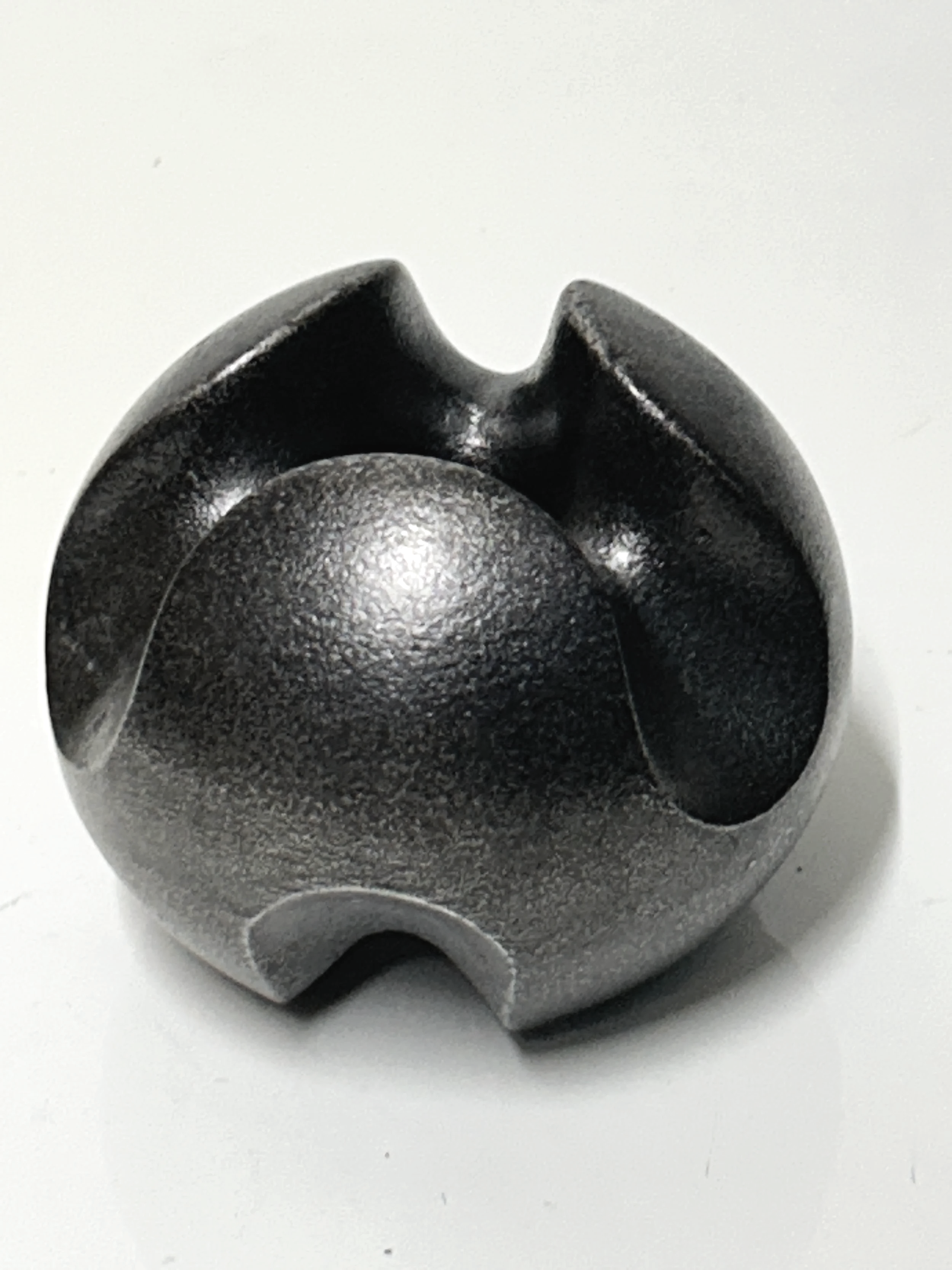 Close-up of a blackened and polished cast iron sculpture, resembling a spherical mechanical part with three evenly spaced notches around its circumference, placed on a white surface.
