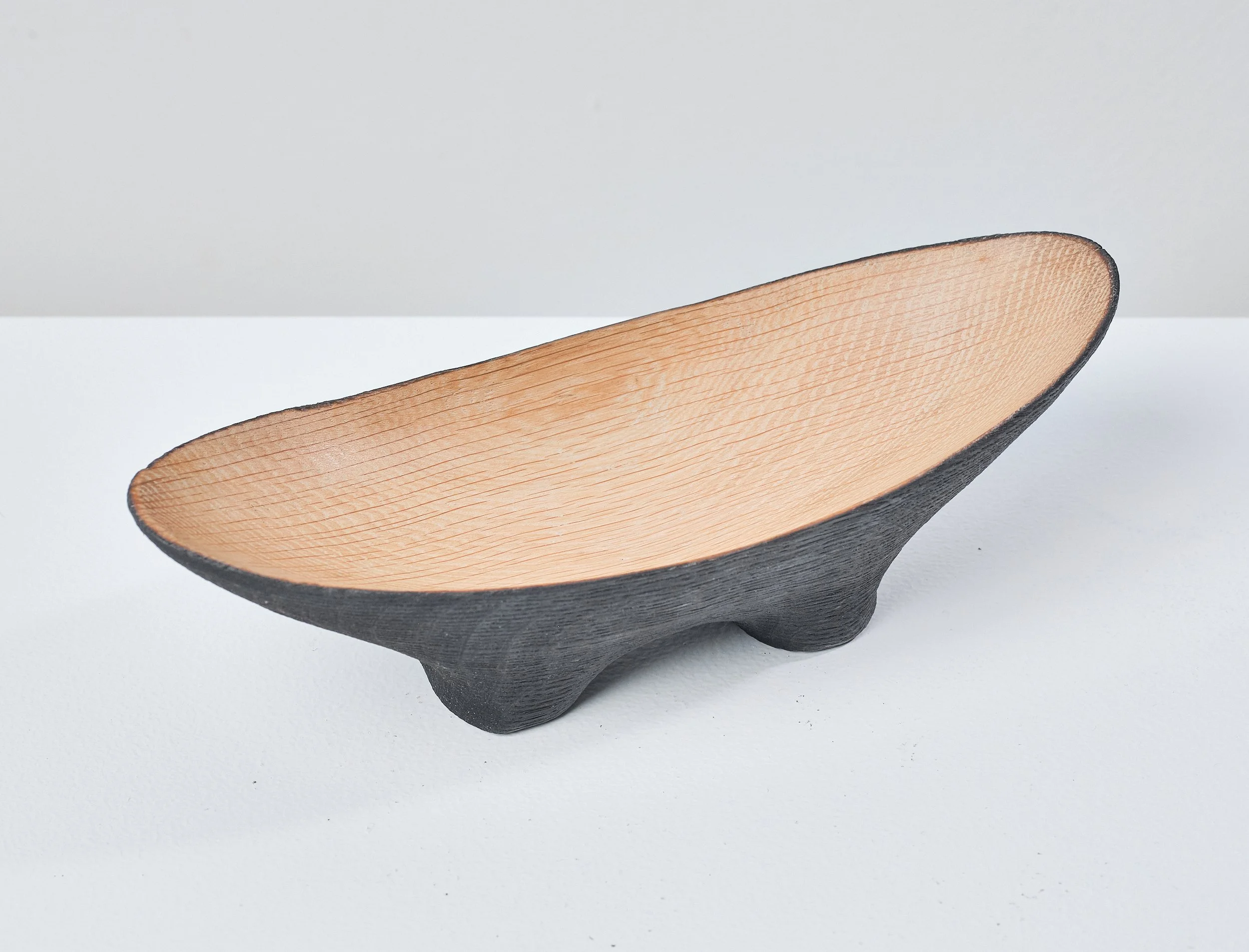 A small wooden bowl made from a salvaged oak beam, with a black exterior and a natural wood interior, placed on a white surface against a plain grey background.
