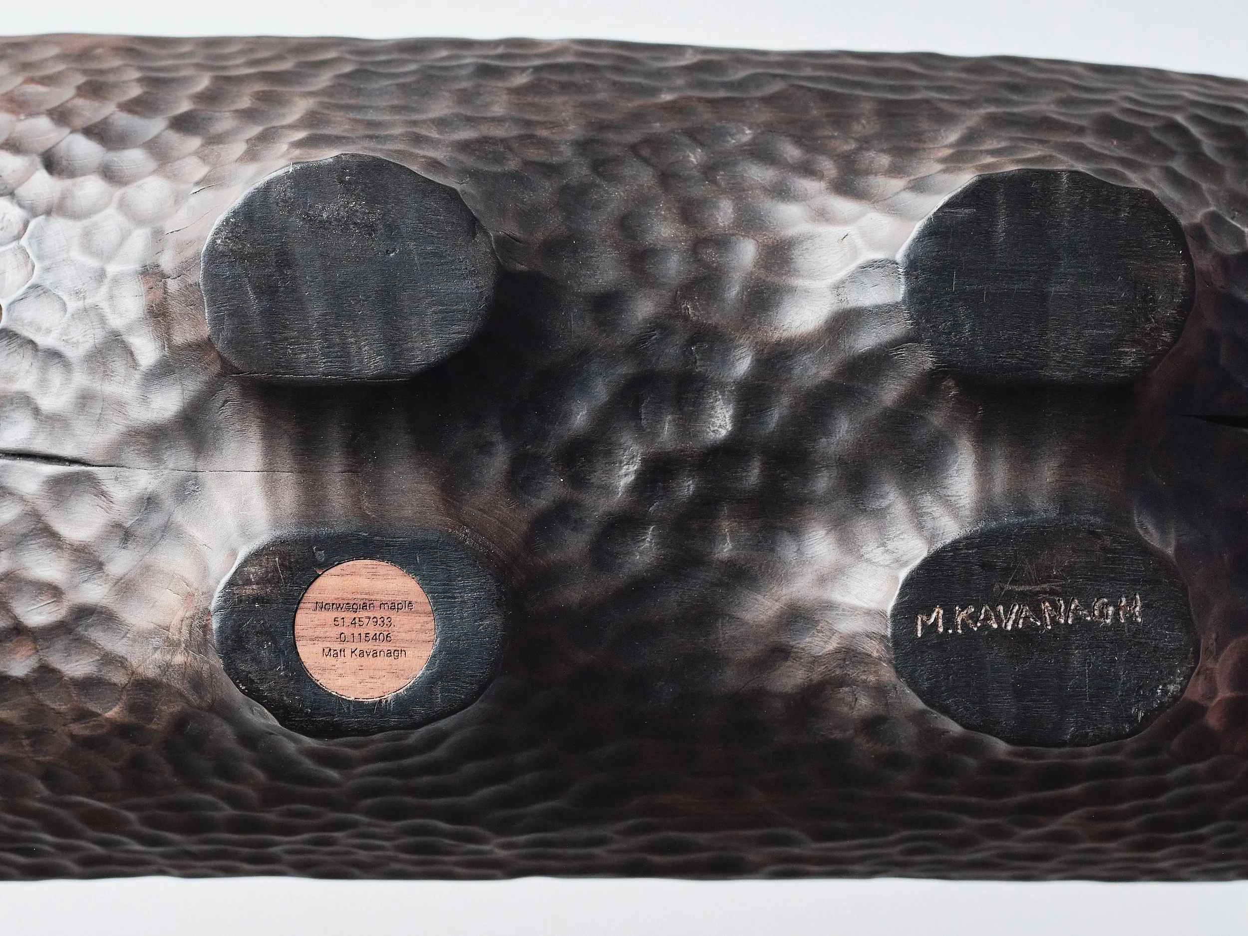 Close-up of the underside of a carved Norwegian Maple wood object with four feet. One foot has a plate with engraved text of the coordinates of where the tree was, sourced the wood species and the artist Matt Kavanagh's name